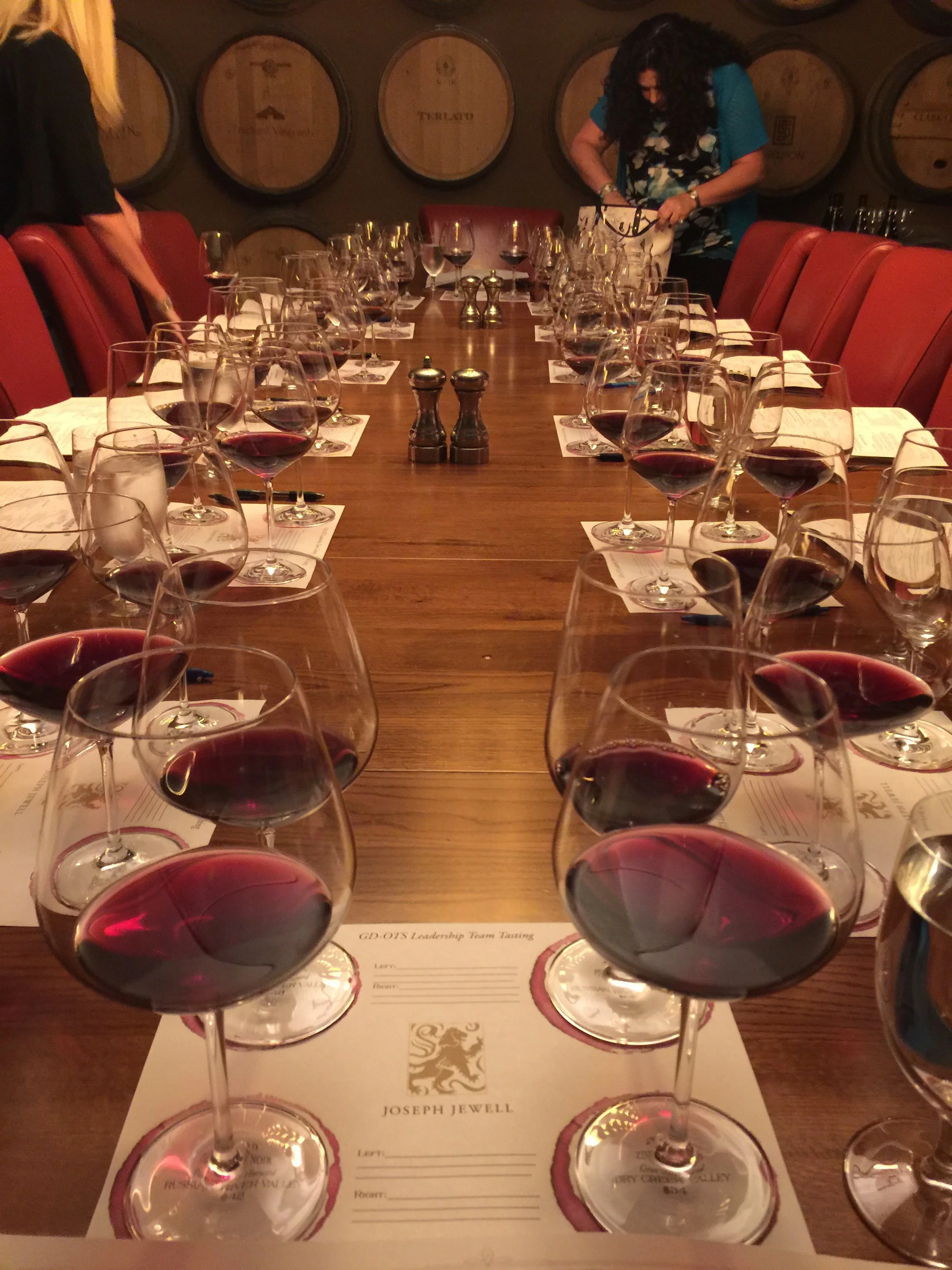 A long wooden table set with glasses of red wine, each placed on a paper with a logo, in a room with wine barrels on the wall. Two women are standing at the far end of the table, preparing for a tasting event.