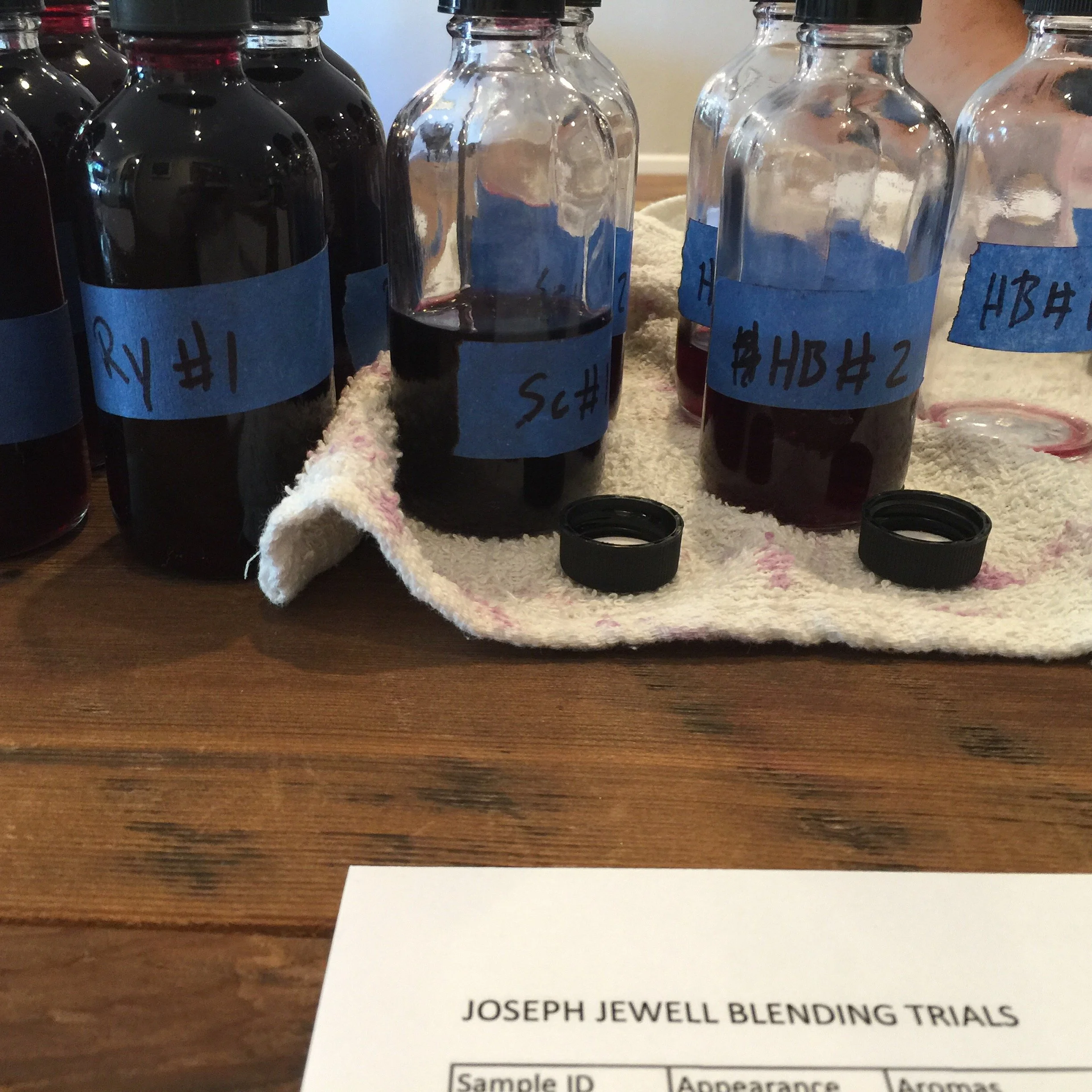 Several small glass bottles filled with a dark liquid, labeled with blue tape and handwritten text, are arranged on a surface. Some of the bottles are empty or partially filled. In the foreground, part of a paper with the title 'Joseph Jewell Blendin