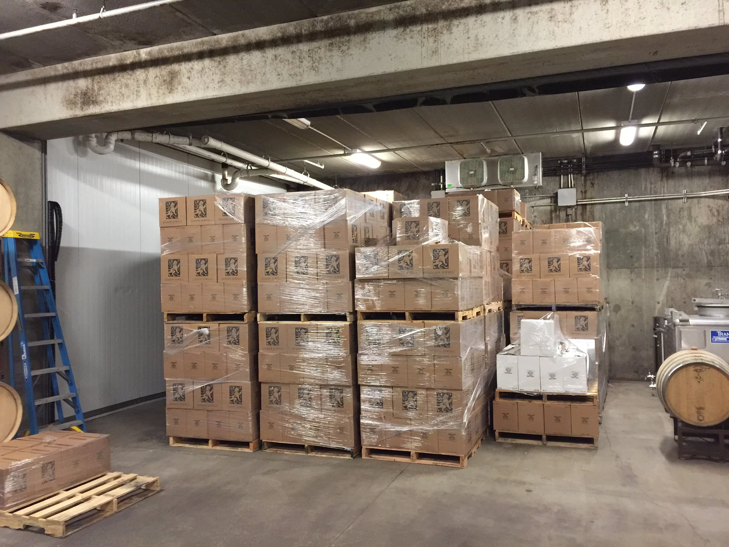 Stacks of cardboard boxes wrapped in plastic on wooden pallets in an underground parking garage or storage area with concrete walls and ceiling, fluorescent lighting, and utility pipes.