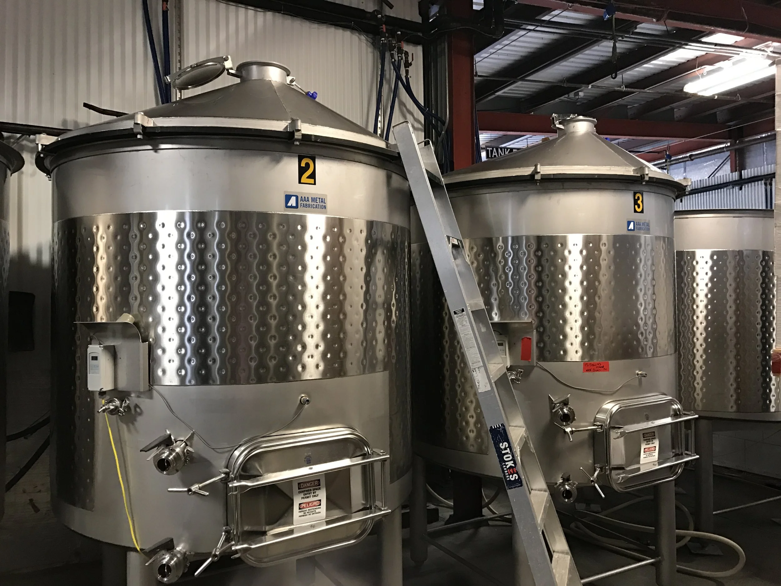 Two large stainless steel tanks with the labels 2 and 3, situated in an industrial setting with pipes, a ladder, and warning signs.
