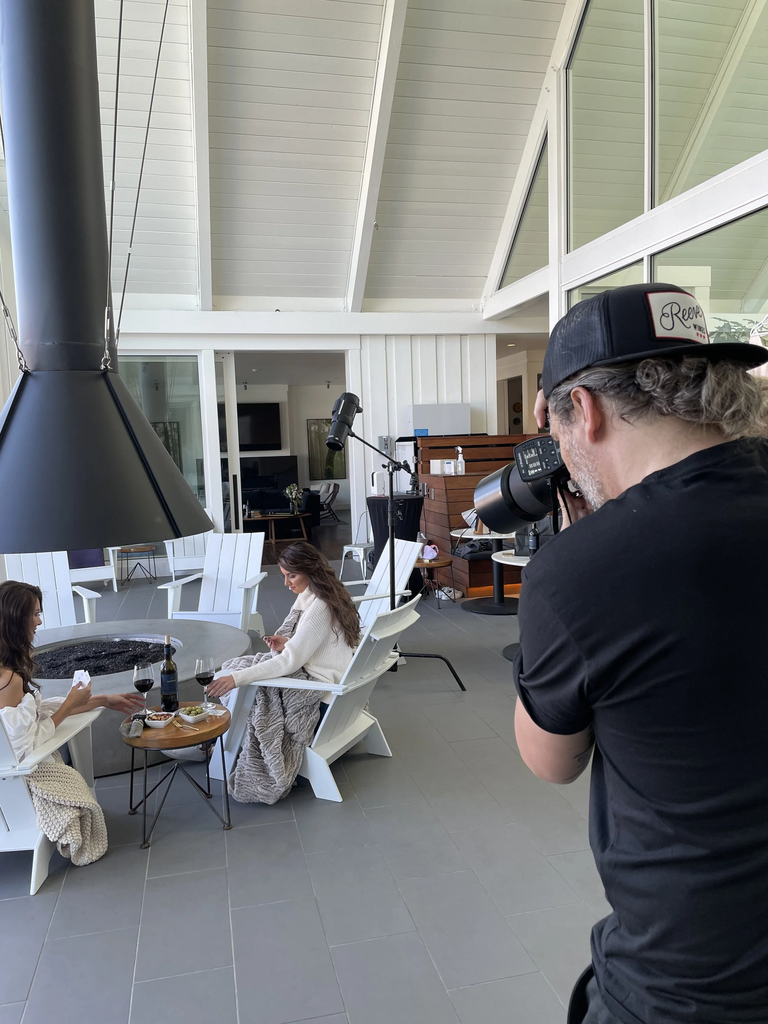 Photographer taking pictures of three women sitting at a table outdoors, with food and drinks, in a modern open space with large windows and a high vaulted ceiling.