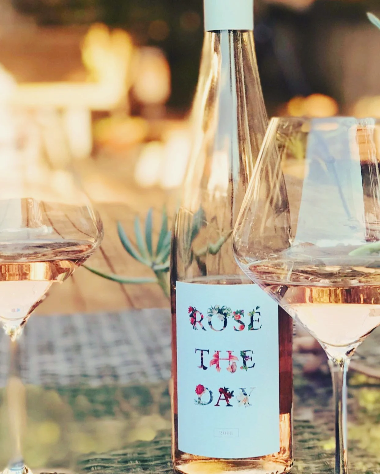 A bottle of rosé wine labeled 'Rose the Day' with two partially visible wine glasses filled with rosé wine placed on a wooden surface, outdoors with blurred warm sunset background.