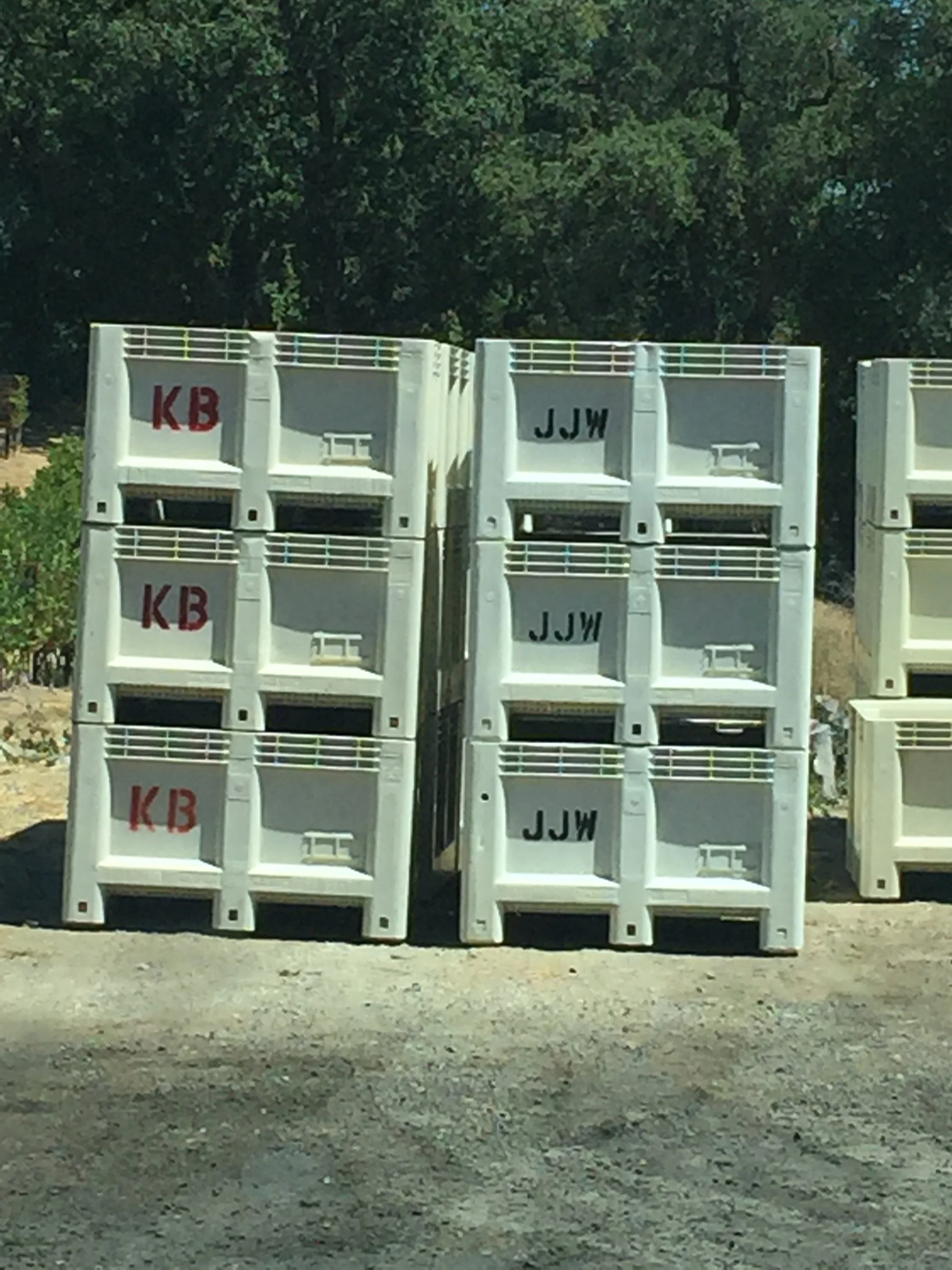 Multiple white plastic crates stacked on the ground outdoors, with some having red and black markings, in front of a background of green trees.