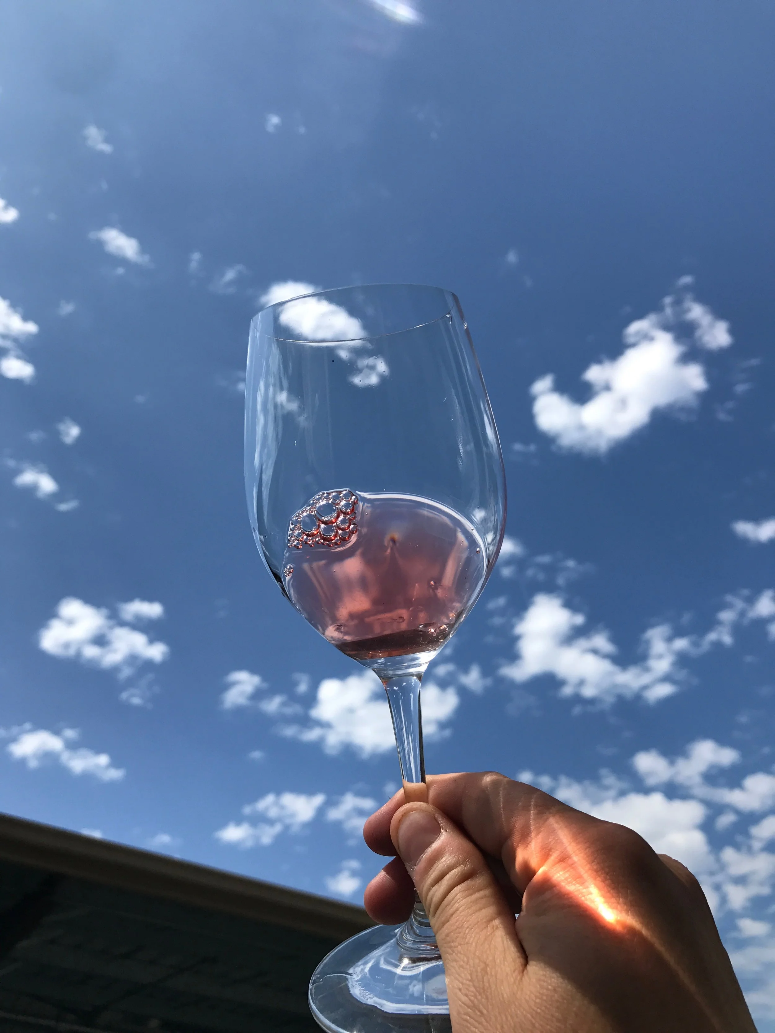 A hand holding a wine glass filled with pink rosé wine against a blue sky with scattered white clouds.