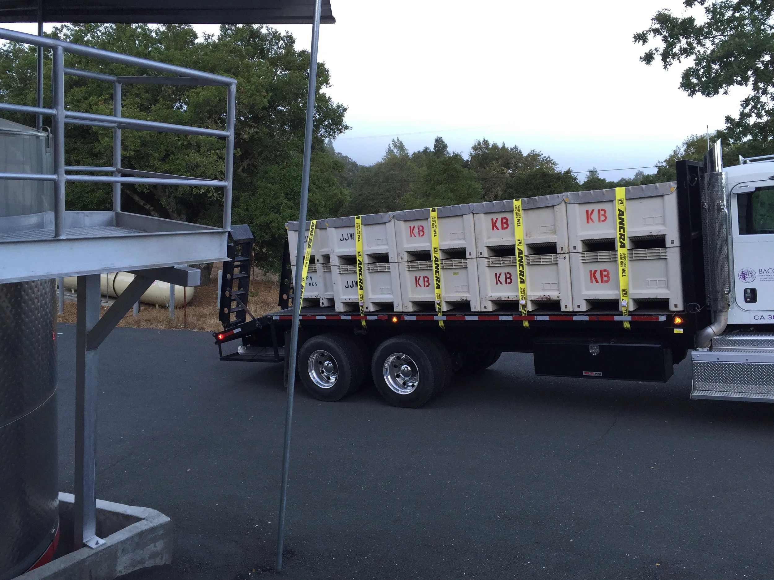 View of a flatbed truck with white crates labeled 'KB' and yellow straps for securing; adjacent metal structure and part of a trailer in the foreground; trees and a cloudy sky in the background.