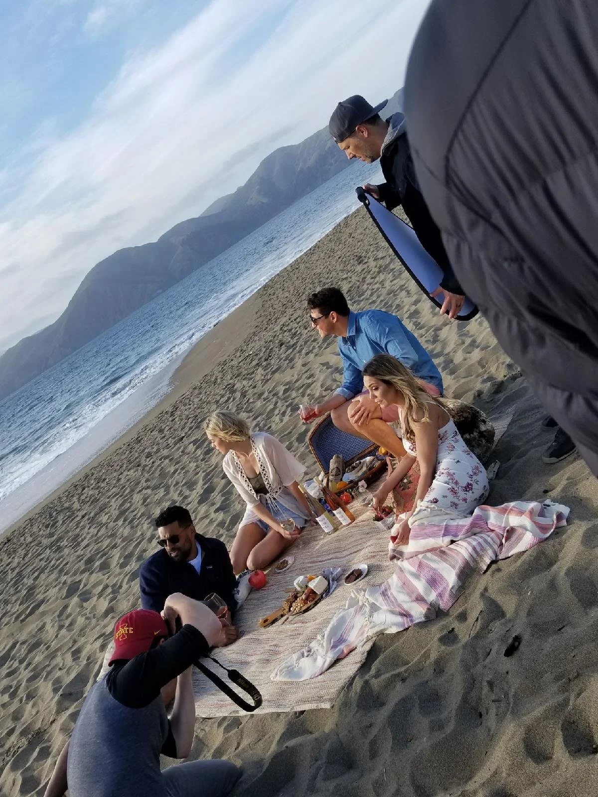 Group of friends having a picnic on the beach with mountains in the background, sitting on a blanket with food and drinks.