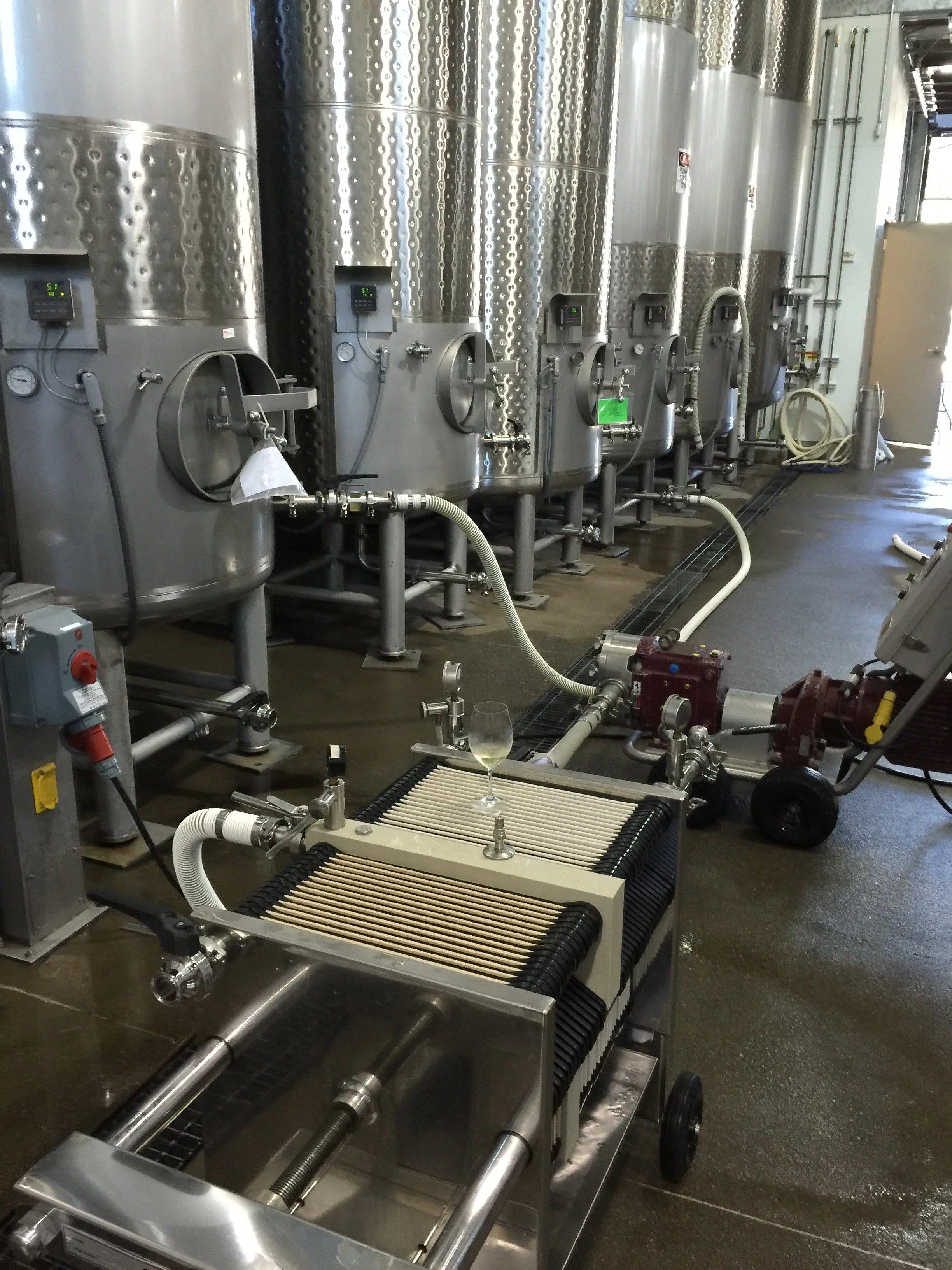 Industrial stainless steel tanks with digital controls in a brewery or winery setting, with hoses and equipment connected for processing liquids.