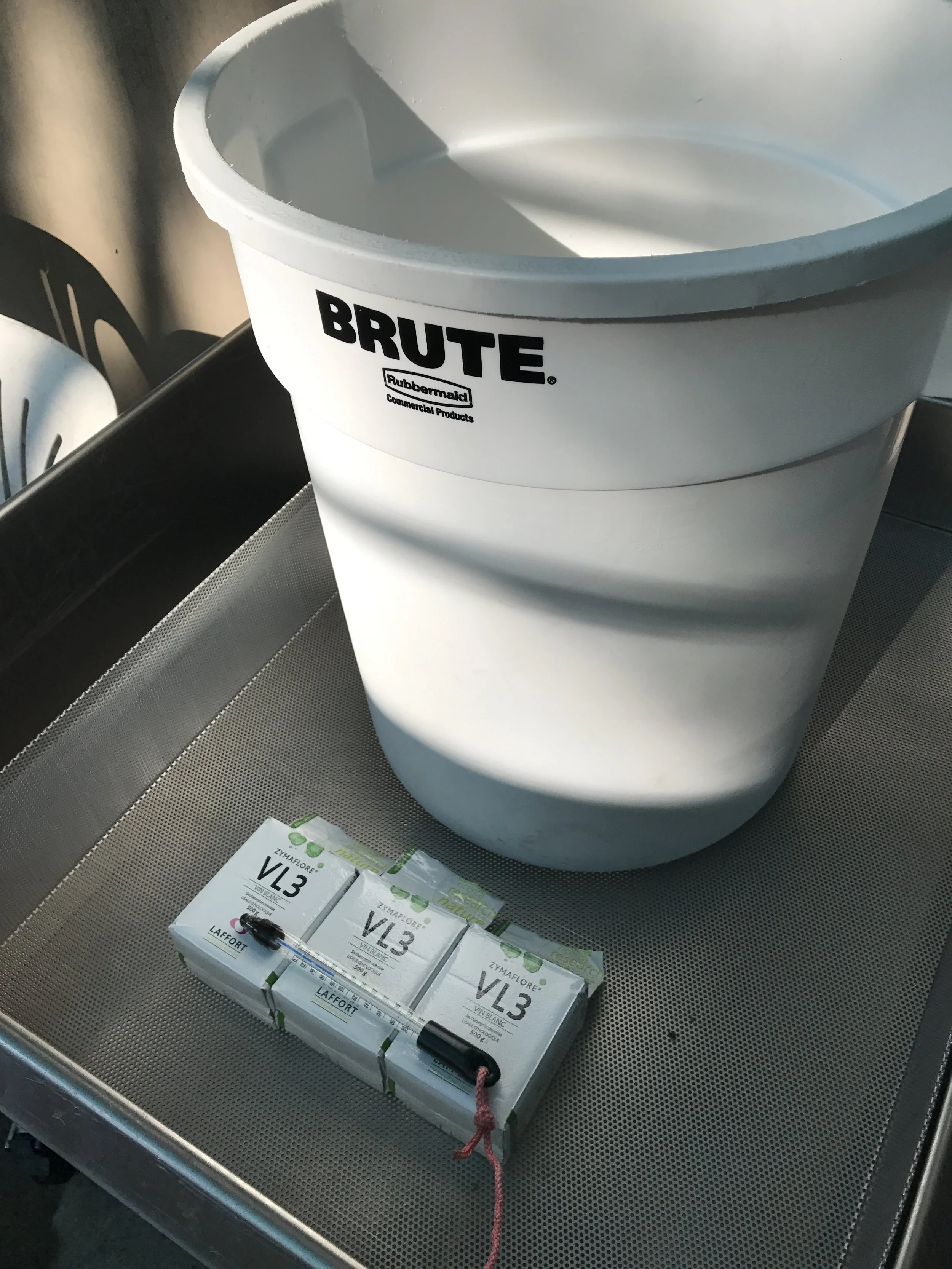 A white bucket labeled 'BRUTE' with 'Rubbermaid' and 'Commercial Products' logos, placed on a metallic surface. In front of the bucket, there are three boxes labeled 'VL3' containing medical or laboratory supplies, and a black marker tied with a pink