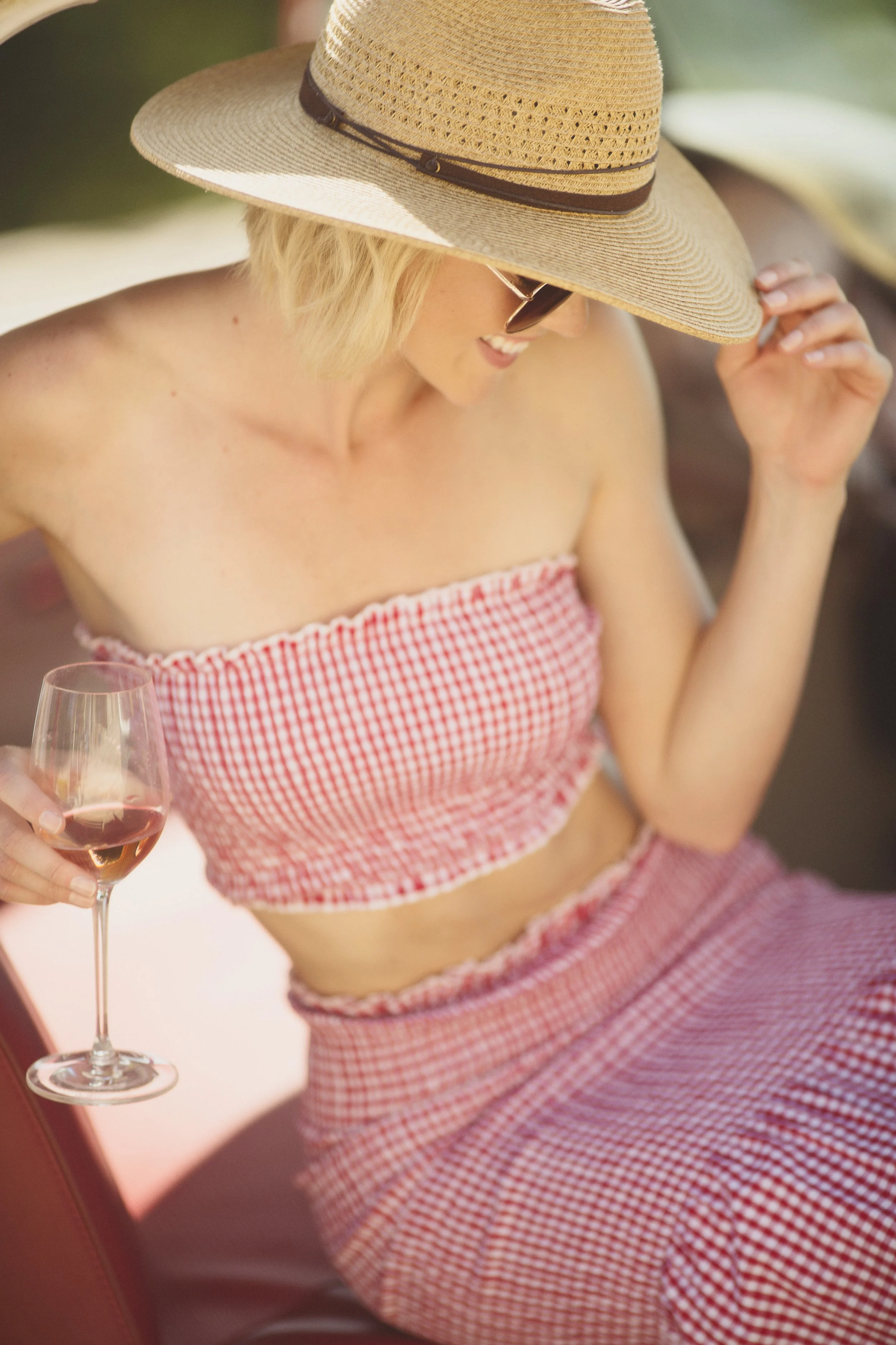 A woman wearing a large straw sun hat with a dark band, sunglasses, and a red and white checkered strapless top, holding a glass of rosé wine.