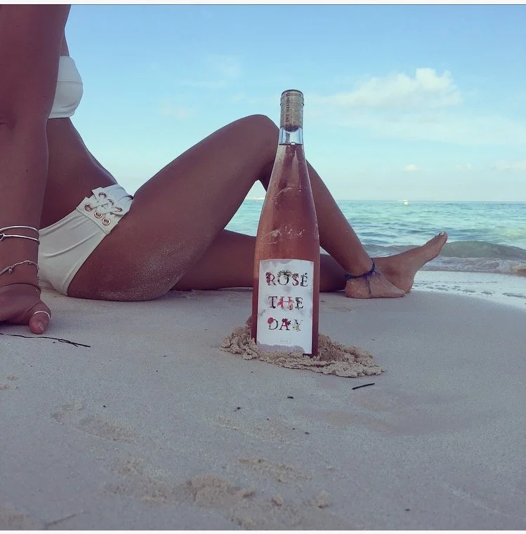 A woman sitting on the beach with her legs extended, nearby is a bottle with a handwritten label saying 'Rose the Day,' partly sunk in the sand, with the ocean and sky in the background.