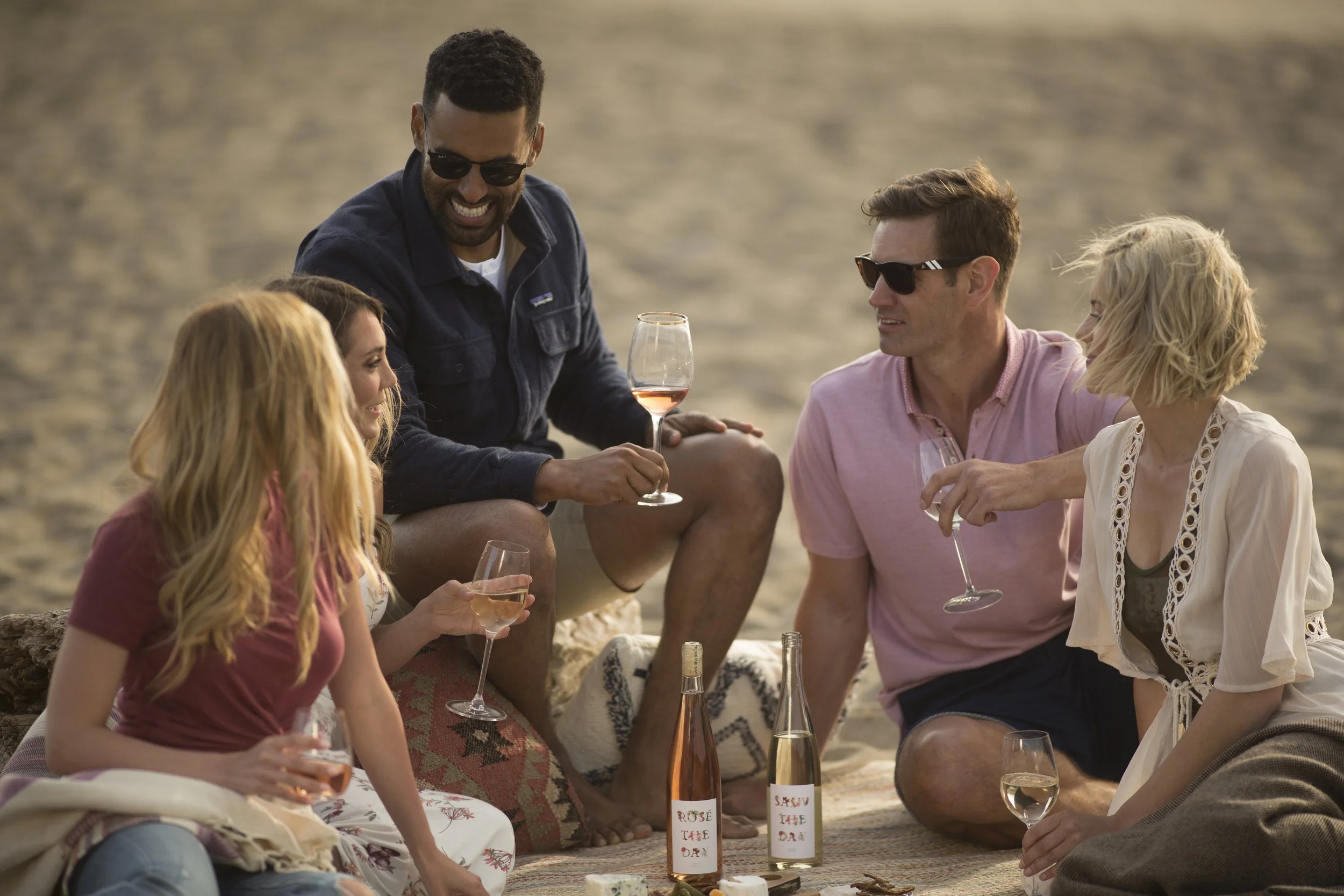 A group of five friends, three women and two men, sitting on a beach, enjoying drinks and conversation, with the ocean in the background during sunset.