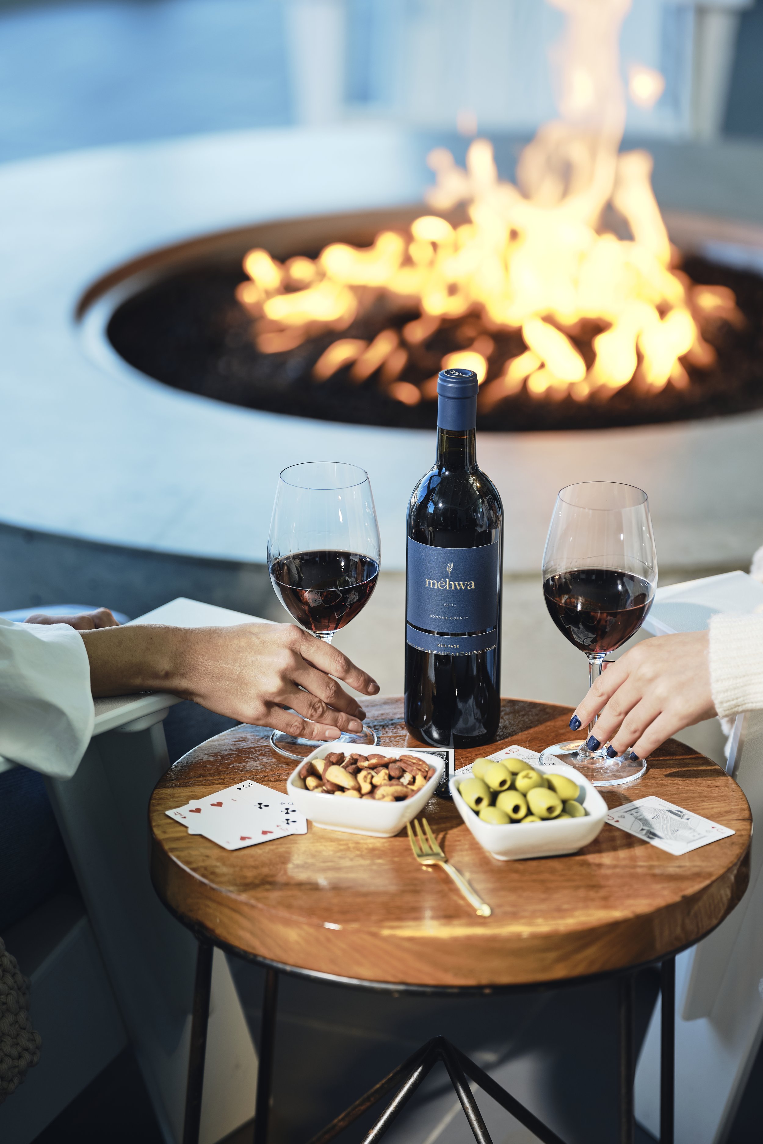 A round wooden table set with two glasses of red wine, a bottle of wine labeled 'méhwa,' bowls of nuts and green olives, playing cards, and a gold fork. In the background, a fire pit with visible flames is present.