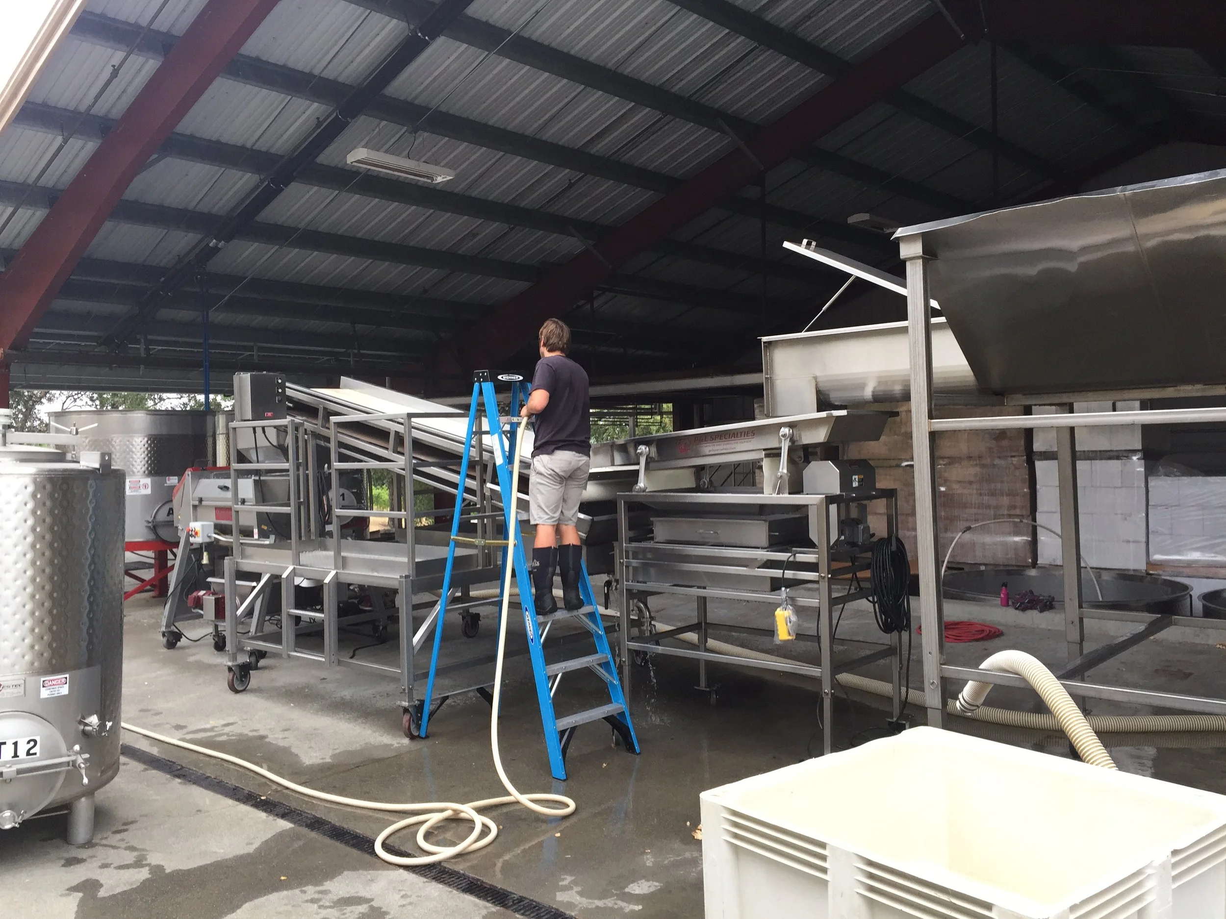 A man standing on a blue step ladder working with stainless steel equipment in an industrial or commercial kitchen setting with large stainless steel tanks and a high ceiling.
