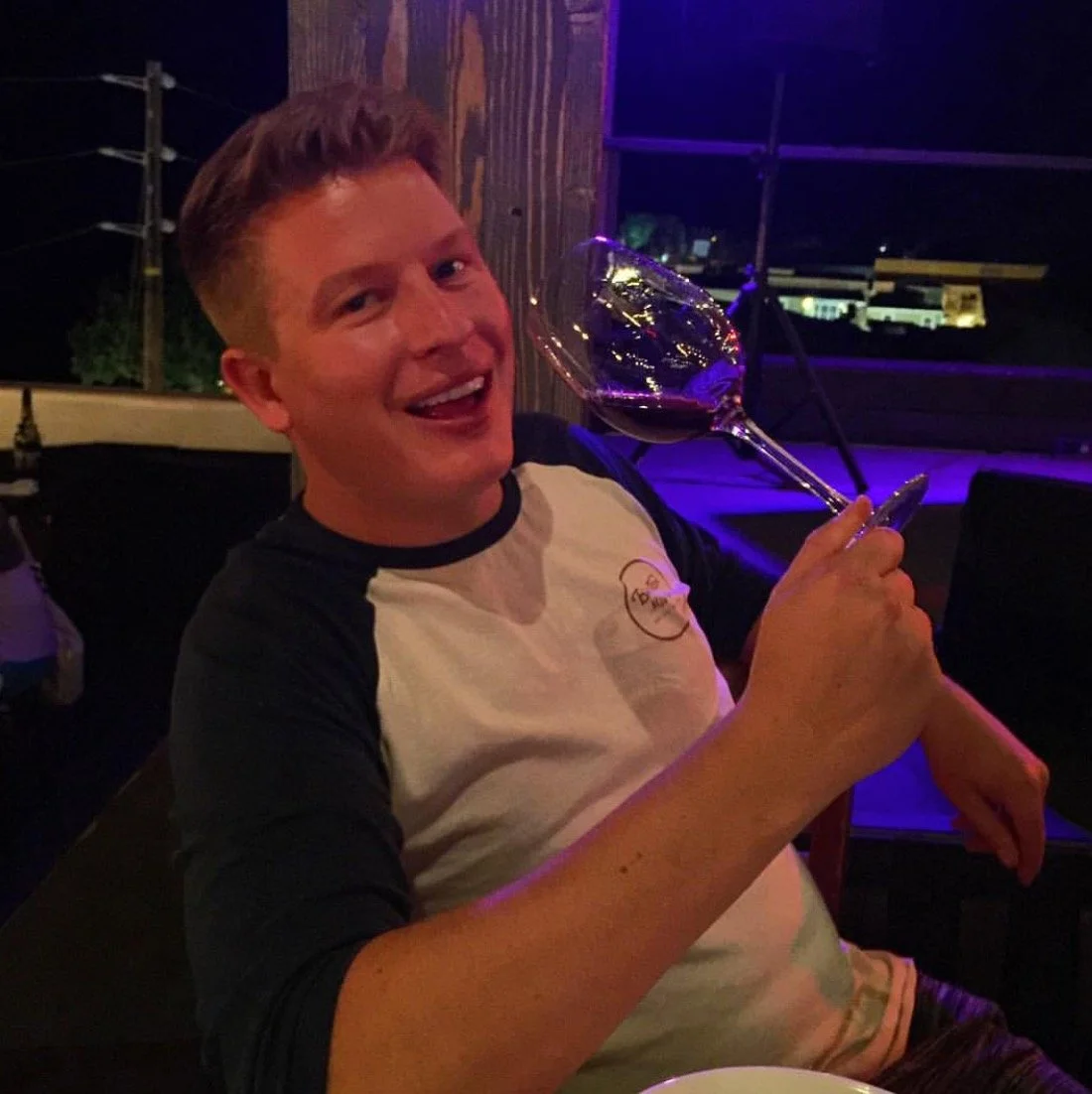 A young man with short hair, smiling and holding a large glass of red wine, sitting at a table in a dimly lit restaurant or bar.