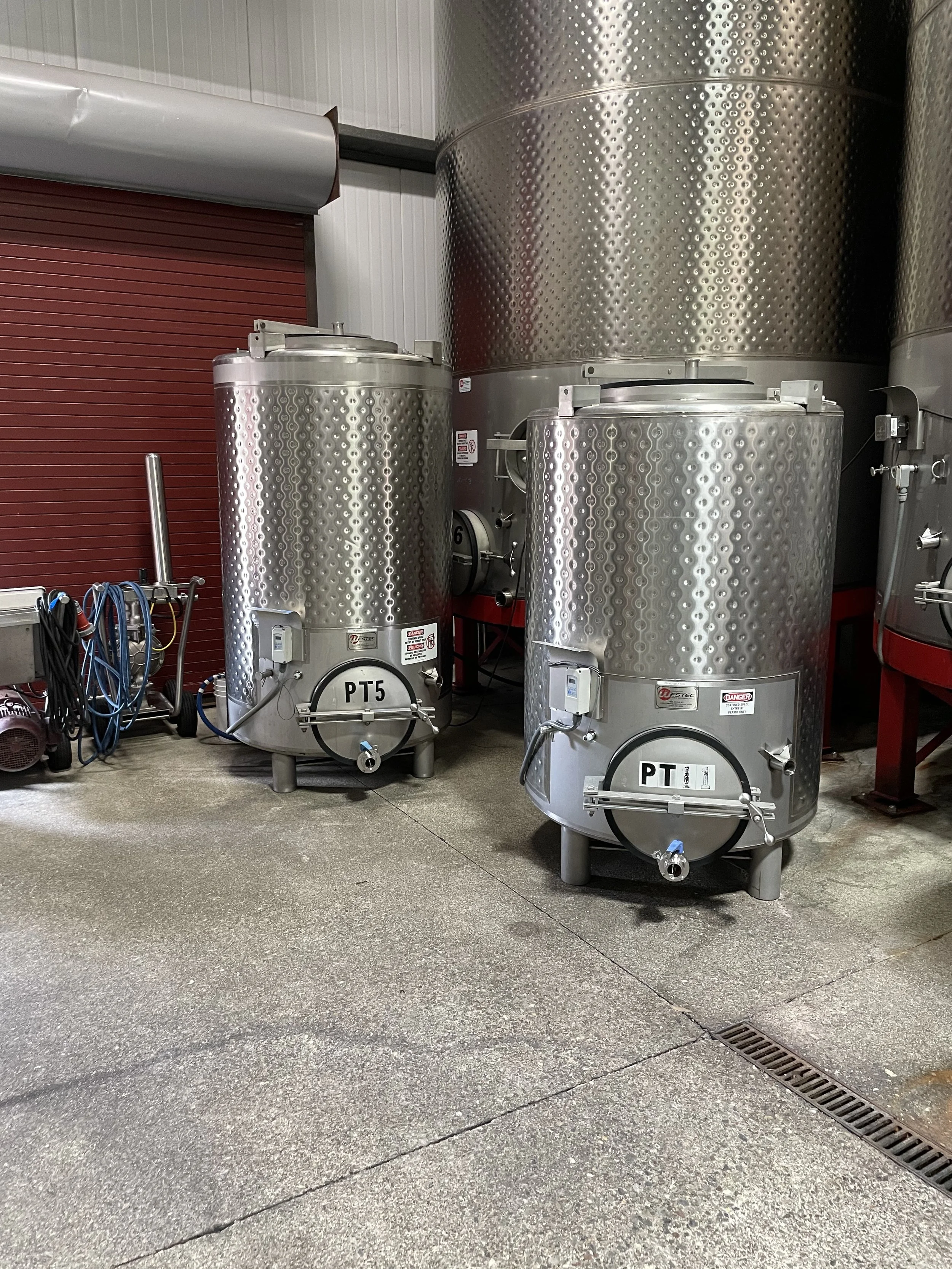 Two large stainless steel fermentation tanks with labels PT5 and PT1 in a brewery or winery setting. There is a red shutter door and some equipment on the left side.