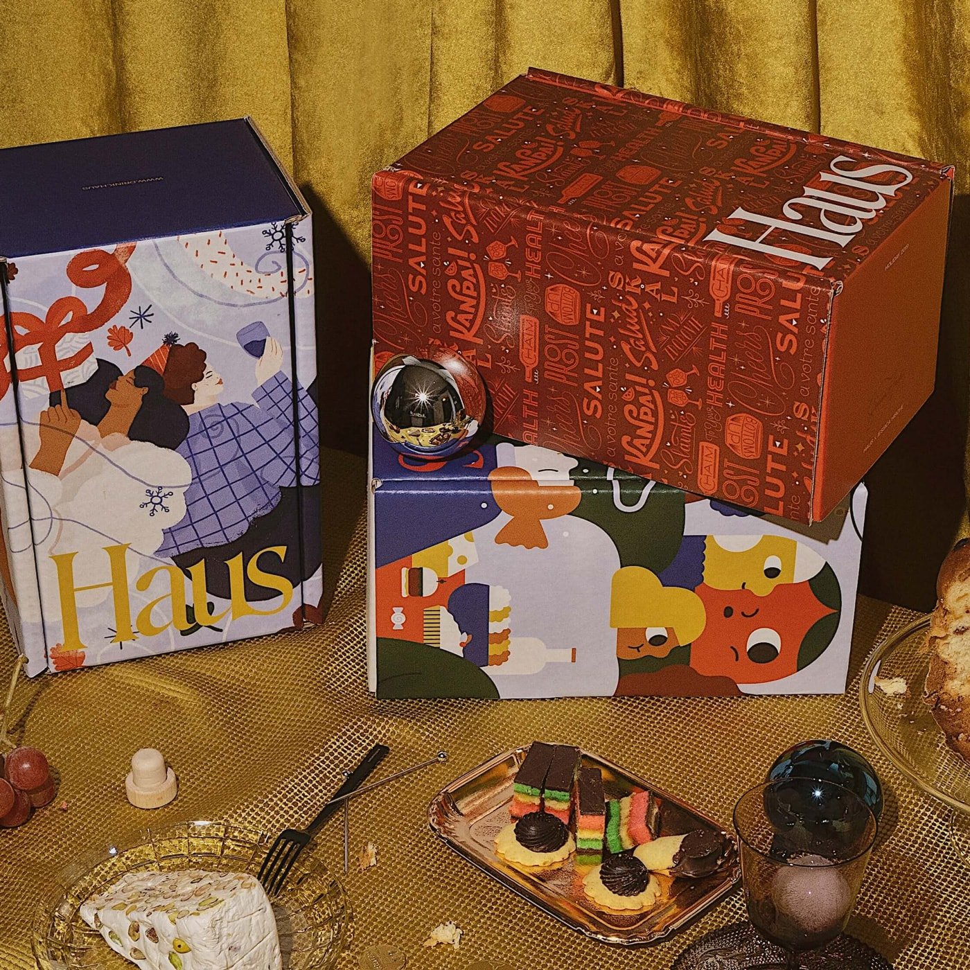 A table with a variety of colorful gift bags, a small cake with layered rainbow colors and chocolate, and a glass of dark soda. The background features a gold curtain.