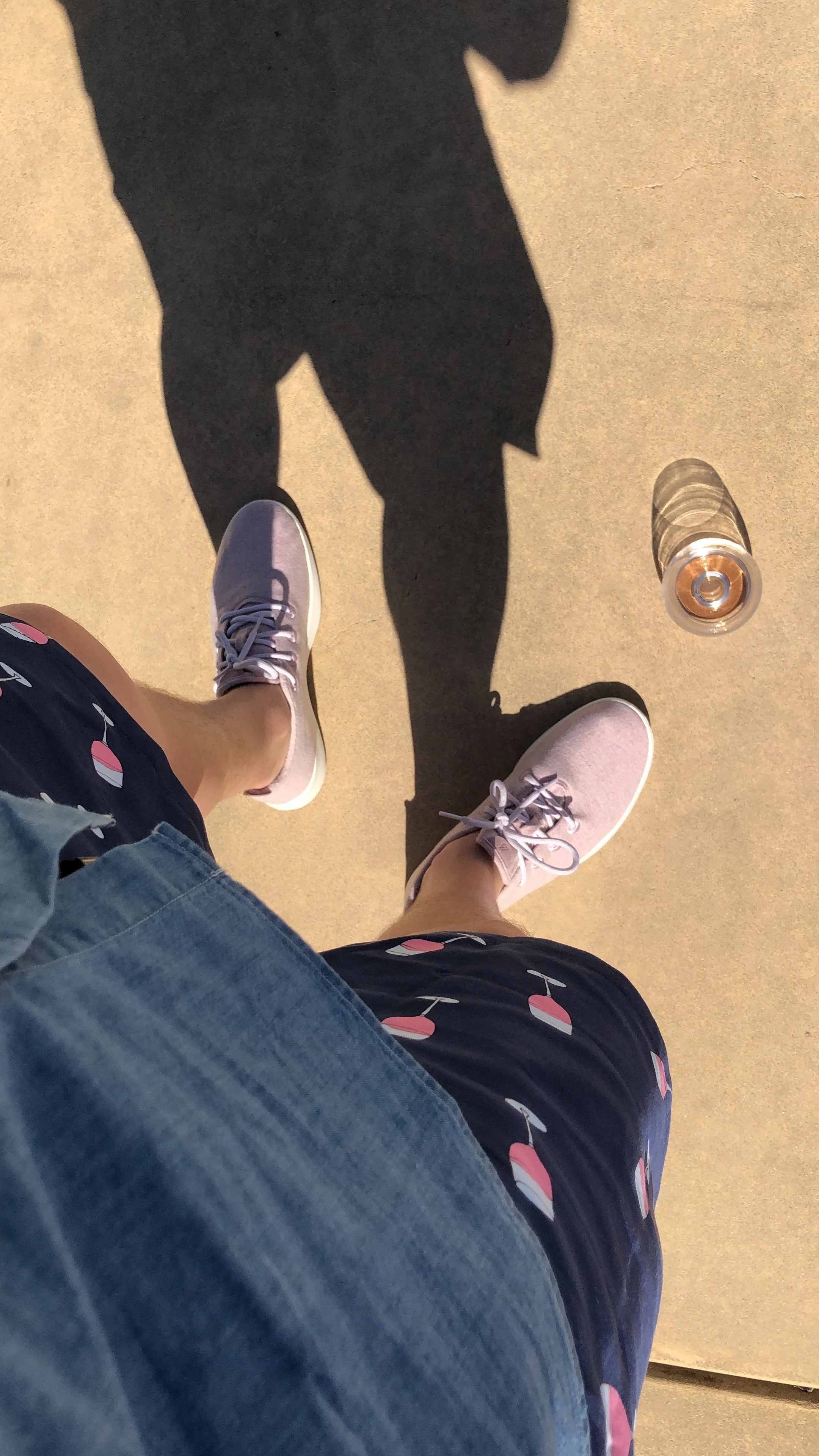 A person's legs and feet wearing pink sneakers, standing on a concrete sidewalk, with a water bottle and a drink on the ground nearby, and their shadow cast on the sidewalk.