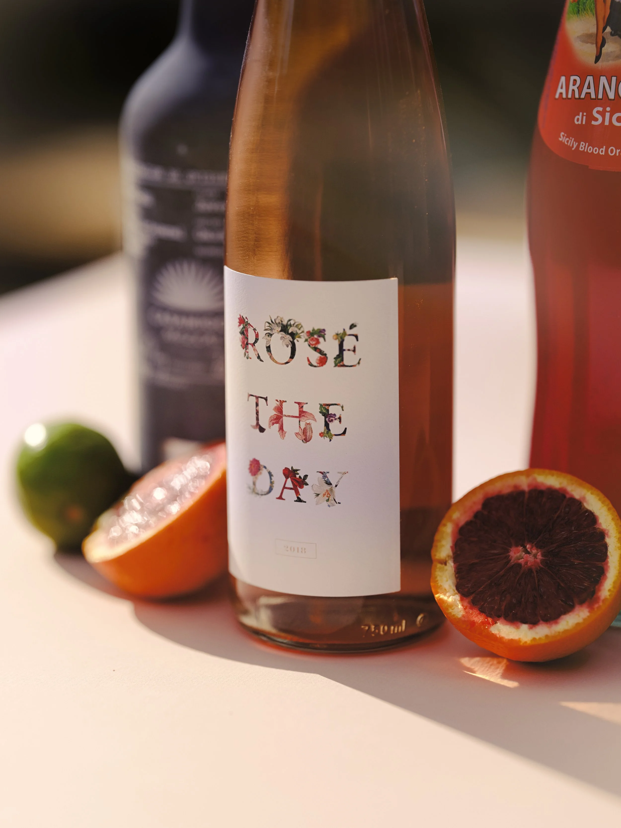 A bottle of rosé wine with a white label surrounded by halved blood oranges, a lime, and a blurred background.