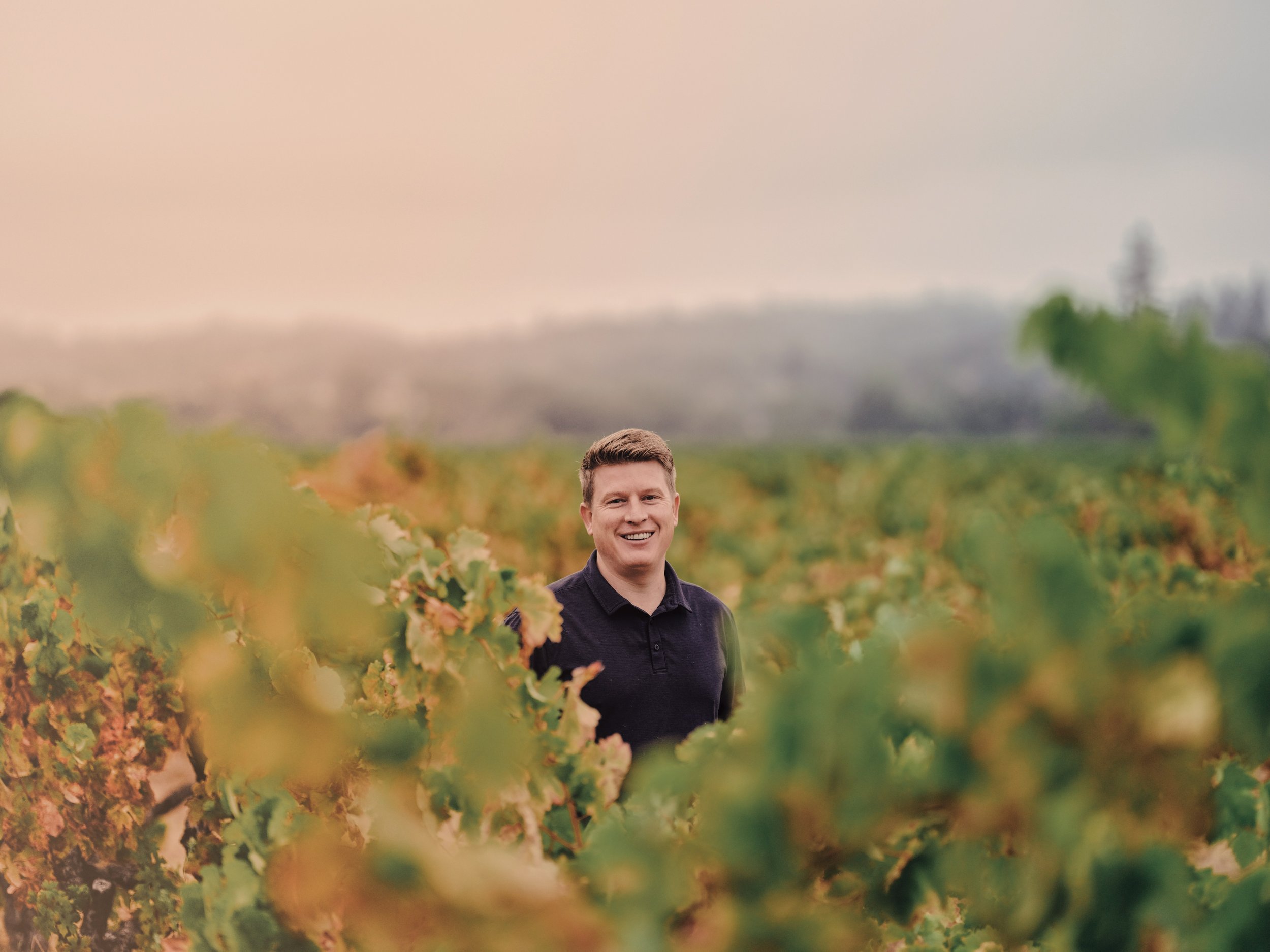 A young man with short brown hair smiling in a vineyard with green and yellow leaves, with blurred trees and a cloudy sky in the background.