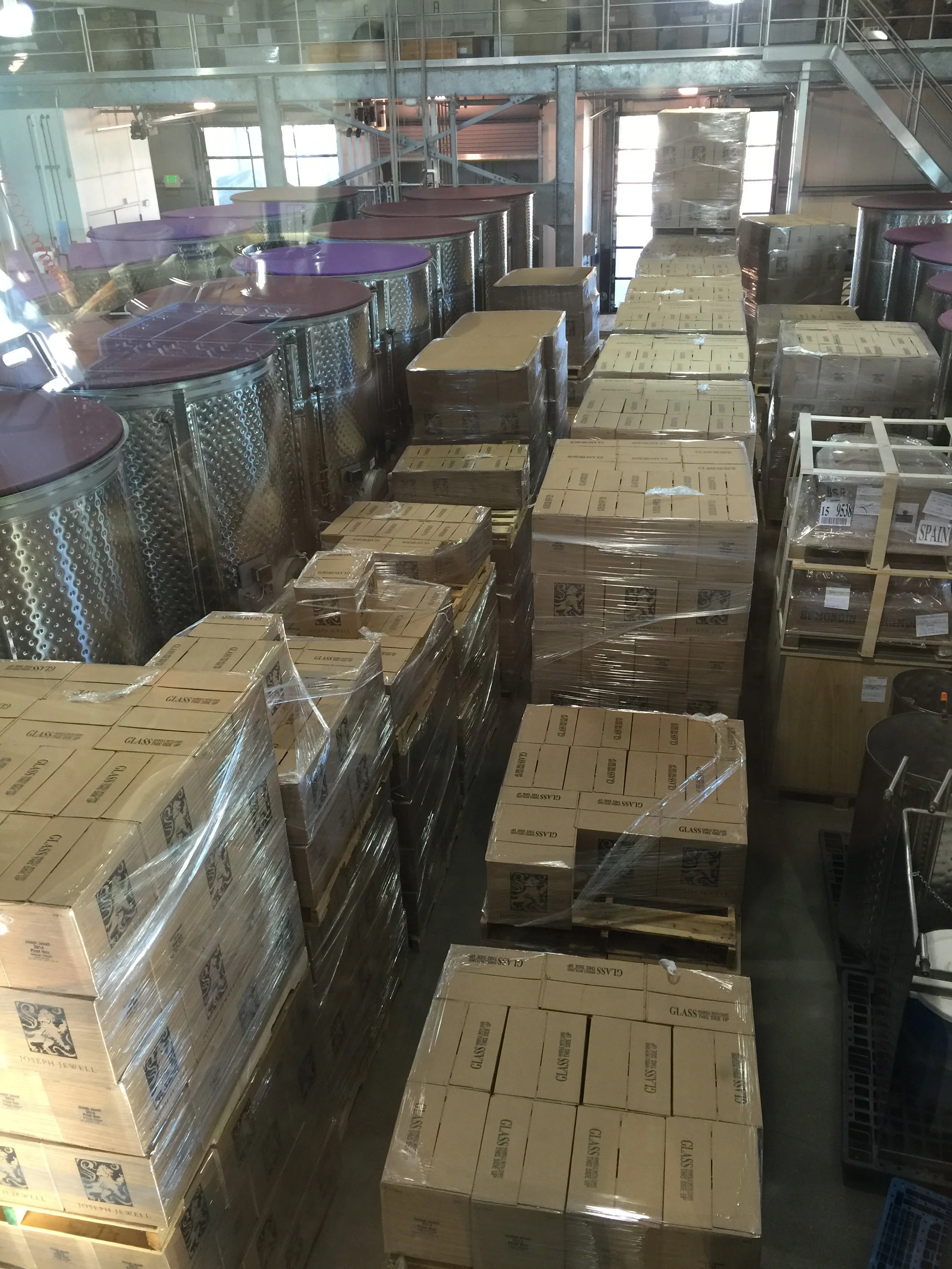 Warehouse with stacked cardboard boxes wrapped in plastic and large metal tanks with purple and brown lids.