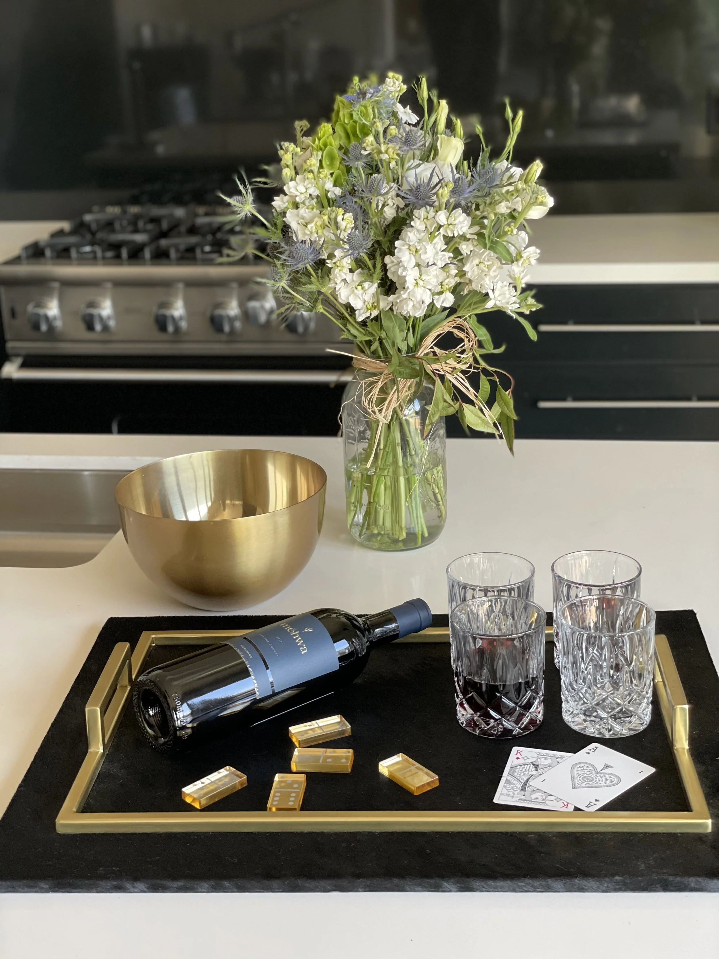 A table with a black surface and a gold border, displaying a wine bottle, four empty glasses, playing cards, and gold domino pieces. In the background, a vase with a bouquet of white and green flowers is visible, with a gold bowl nearby and a black s