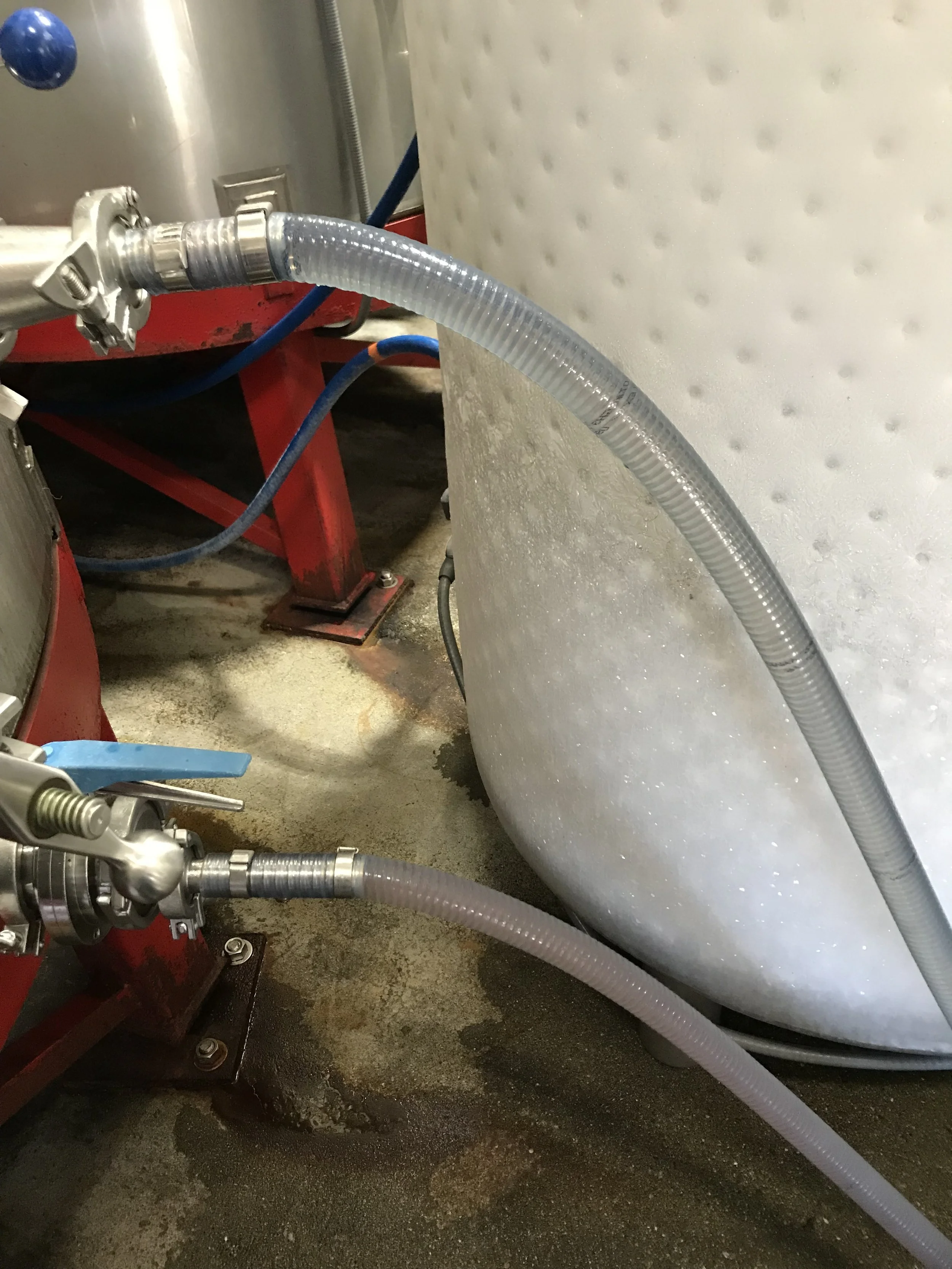 Close-up of industrial piping and hoses connected to a large metal tank or vessel, with wet floor underneath.