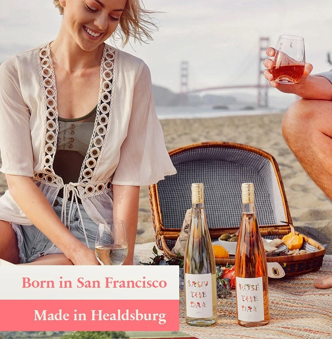 A woman enjoying a picnic on the beach with a bottle of white wine, another bottle of rosé wine, and a glass of rosé in hand. A wicker picnic basket is open on a blanket with food, and the Golden Gate Bridge is visible in the background. Text on the 