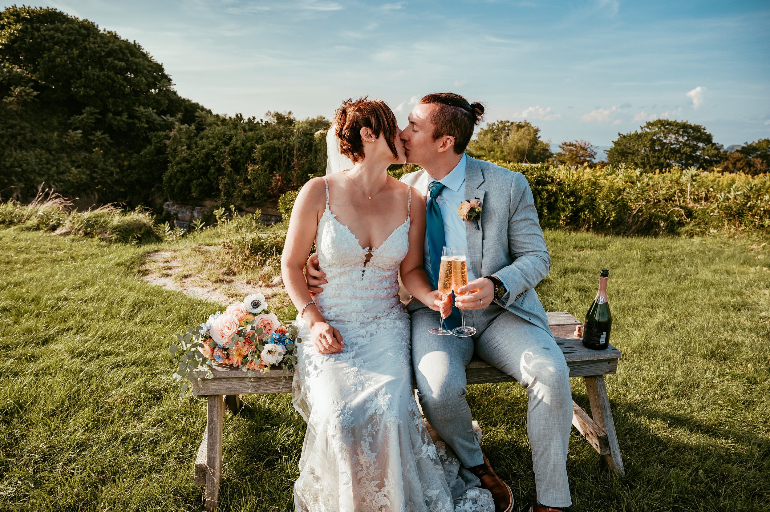 A newlywed couple in wedding attire sharing a kiss and holding glasses of champagne while sitting on a wooden bench outdoors during daytime. The bride is in a white lace wedding dress with a bouquet beside her; the groom is in a light grey suit with 