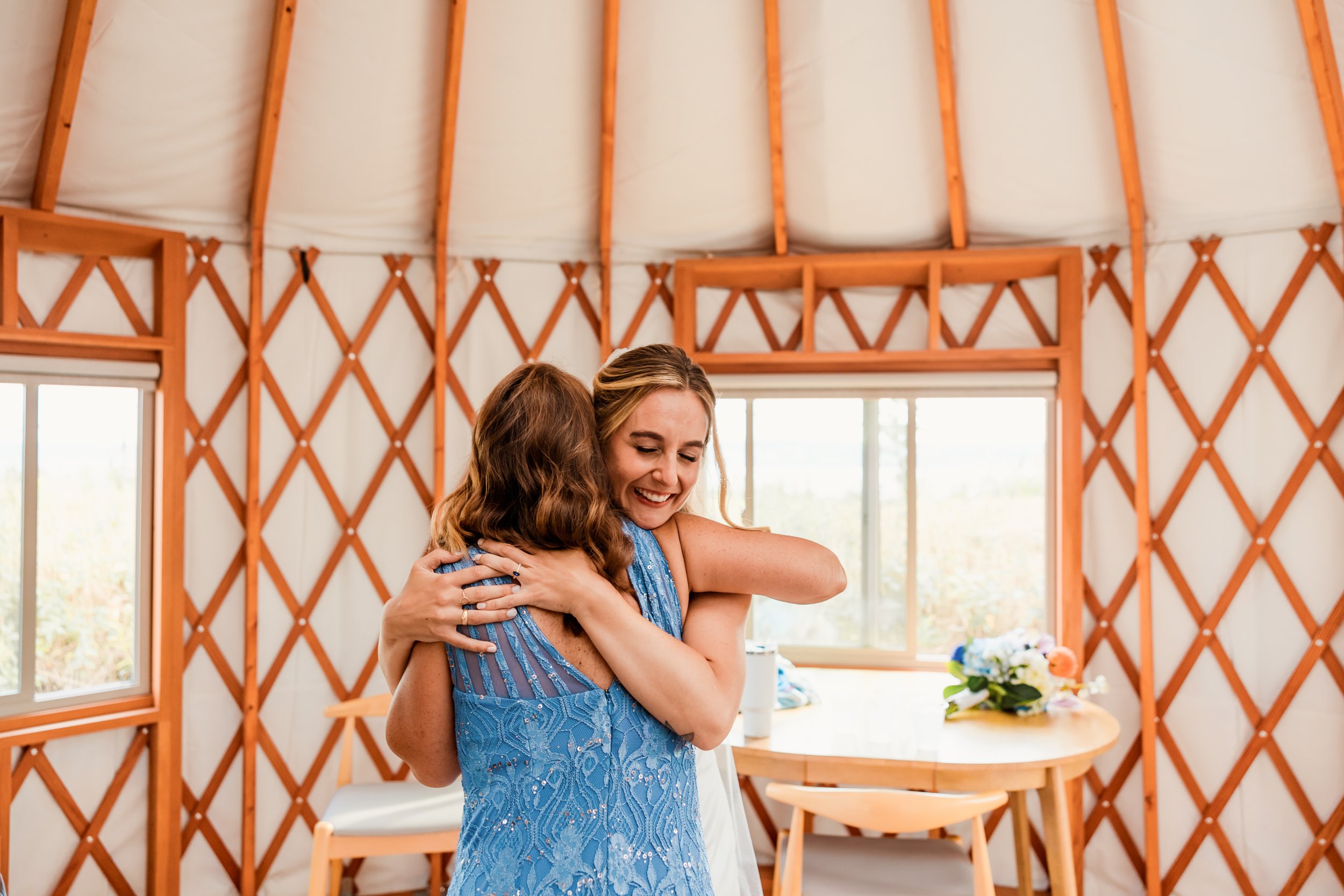 Two women hugging and smiling inside a wooden yurt with large windows and a table with flowers in the background.