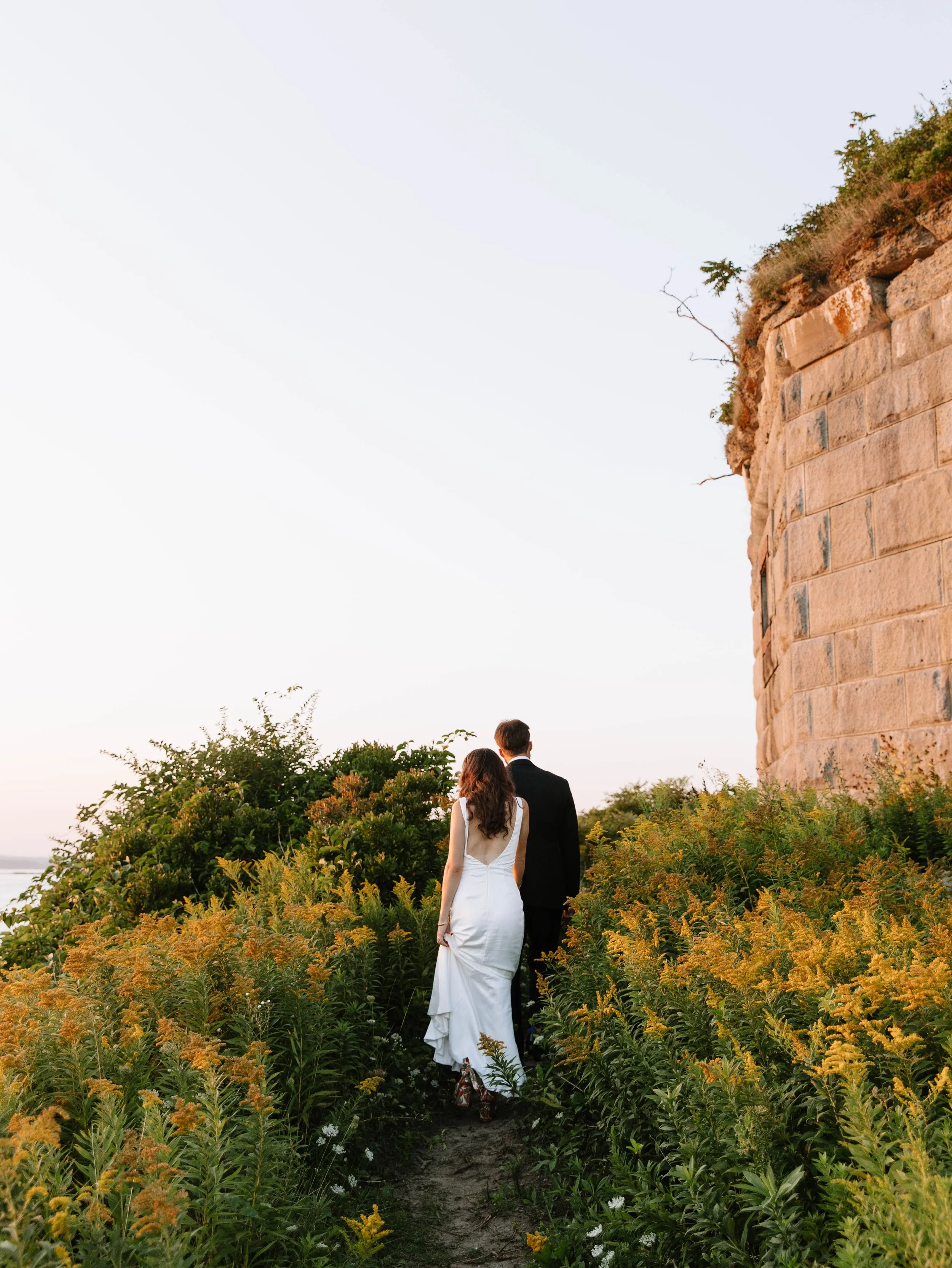 A bride and groom walking down a narrow dirt path surrounded by yellow and green foliage, near a large stone structure, during sunset.