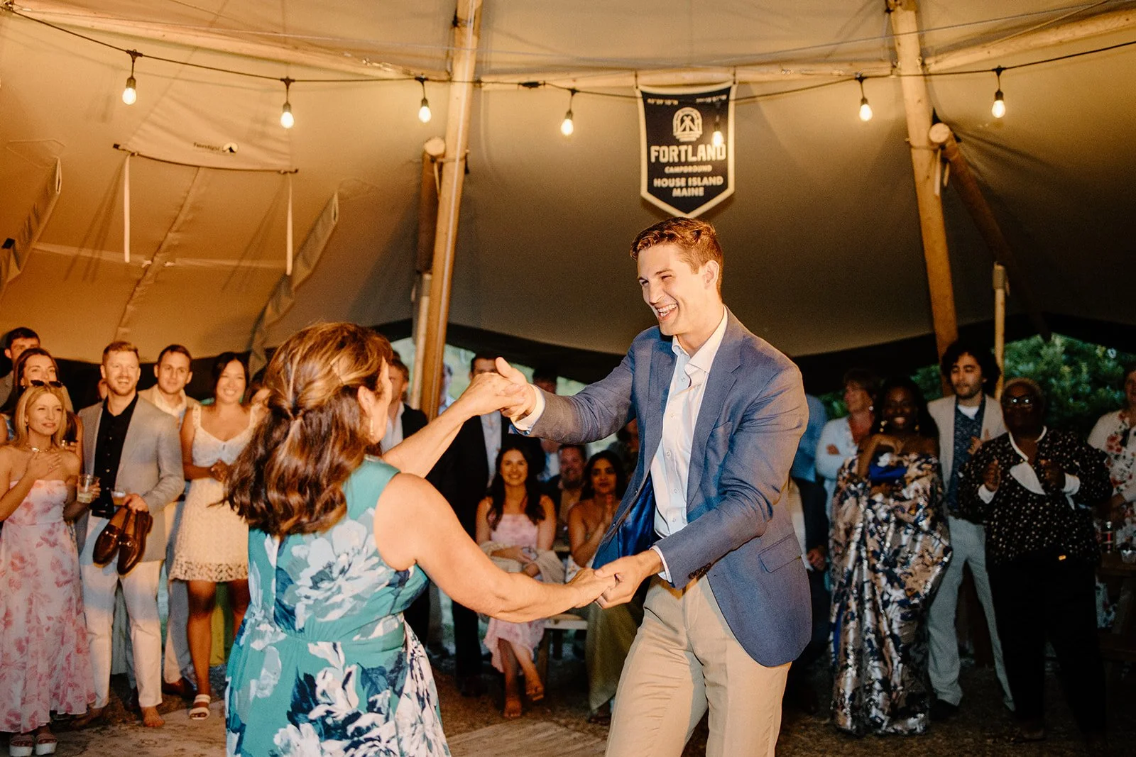 A young man in a blue blazer and beige pants dancing and smiling with an older woman in a floral dress during a celebration under a large tent, with several people watching and enjoying the event.