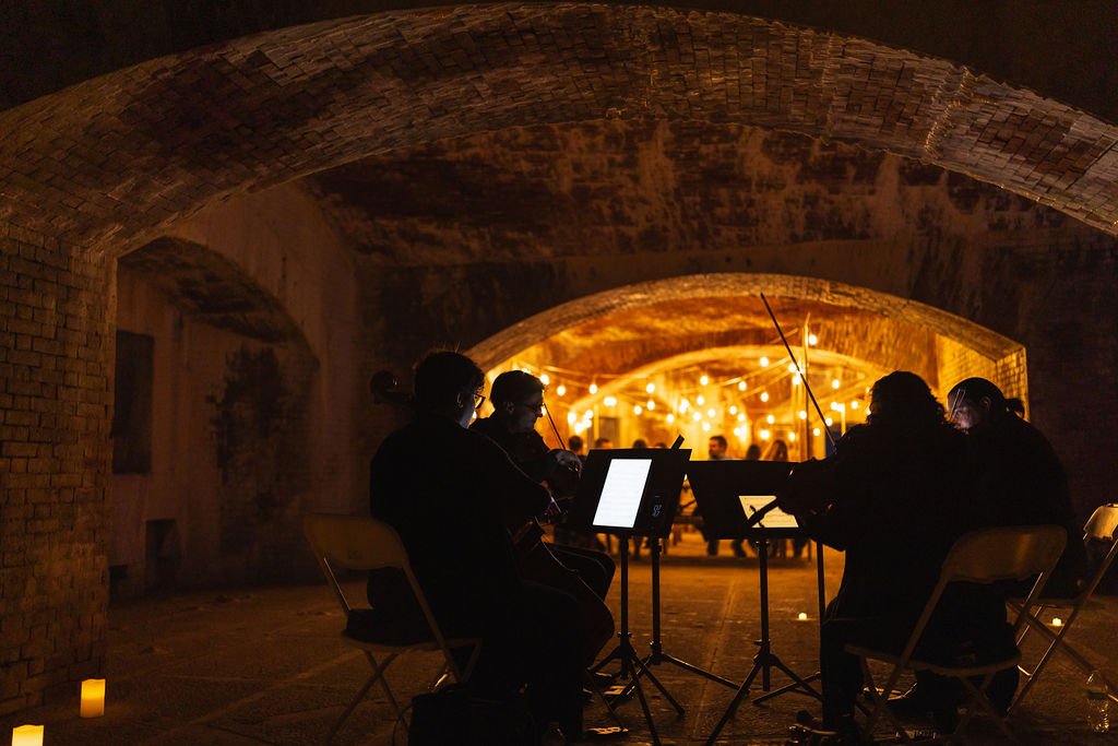 A group of musicians playing string instruments under a brick archway illuminated with warm string lights, with an audience sitting in the background.