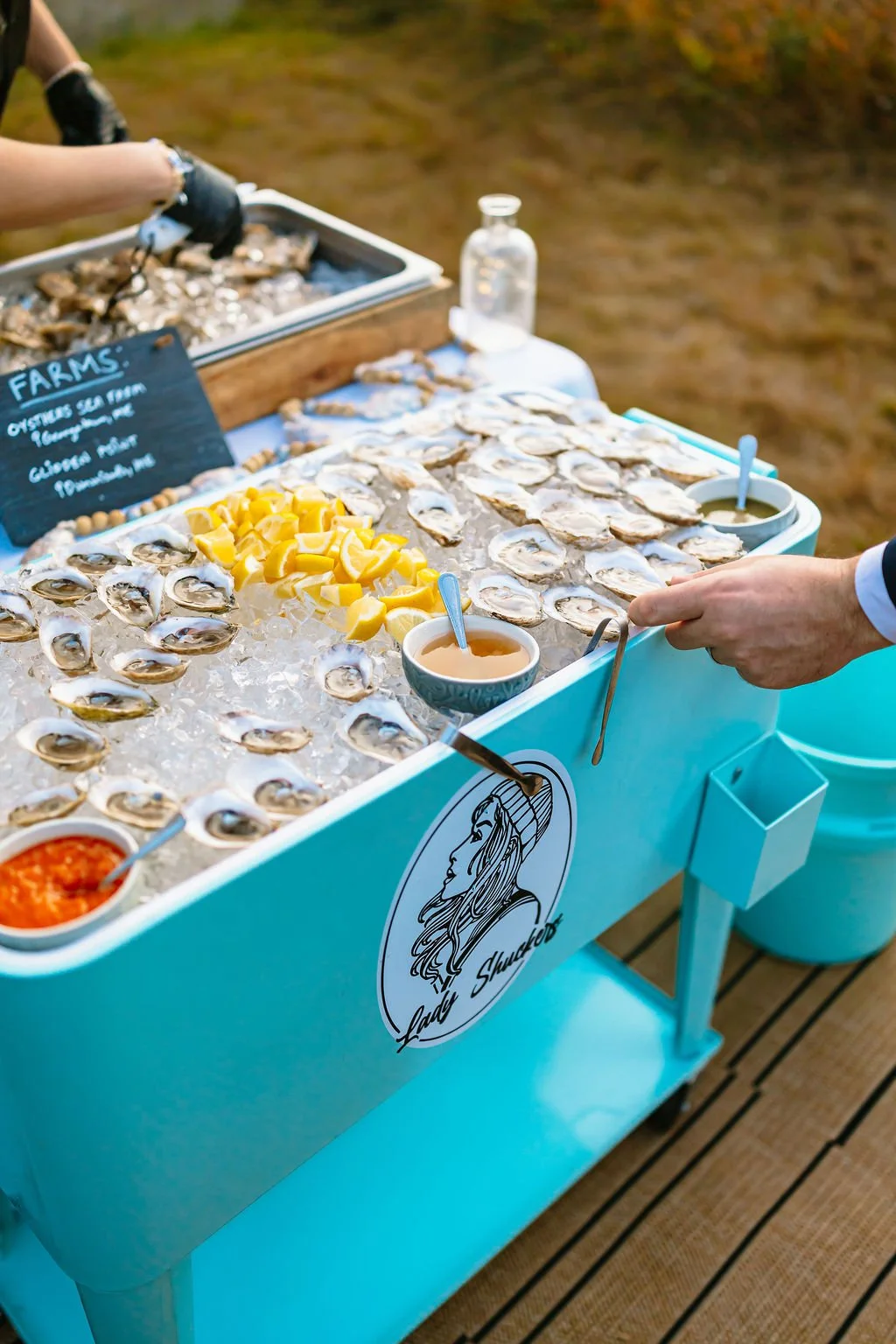 A seafood display at a farm market, featuring fresh oysters on ice, lemon wedges, sauce, and a person using tongs to pick up an oyster.