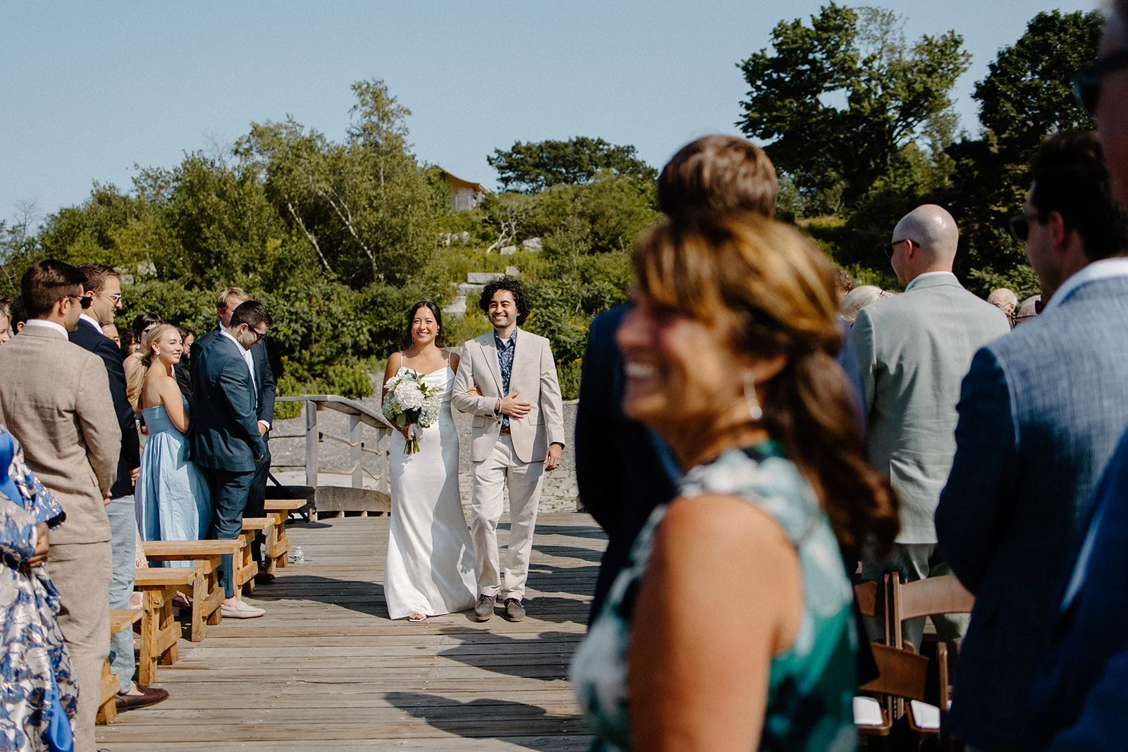 A bride and groom walking down the aisle during their outdoor wedding ceremony on a sunny day, surrounded by seated guests and lush green trees.