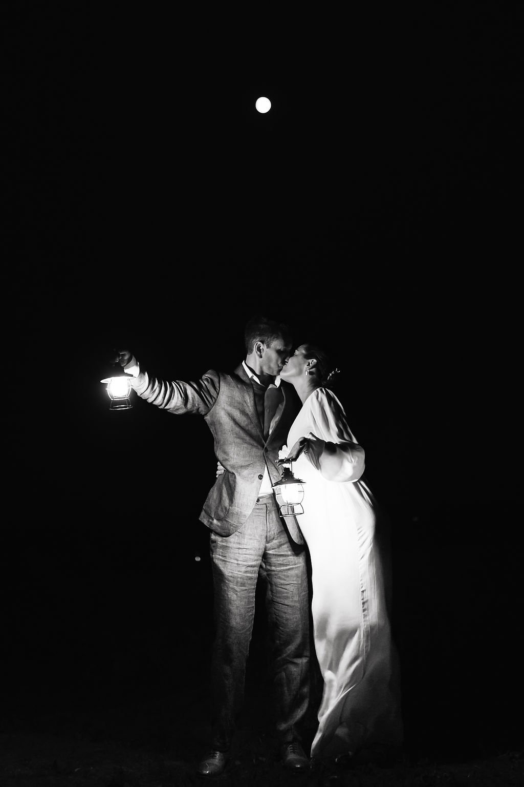 Black and white photo of a couple kissing at night, holding lanterns, with the moon in the sky.