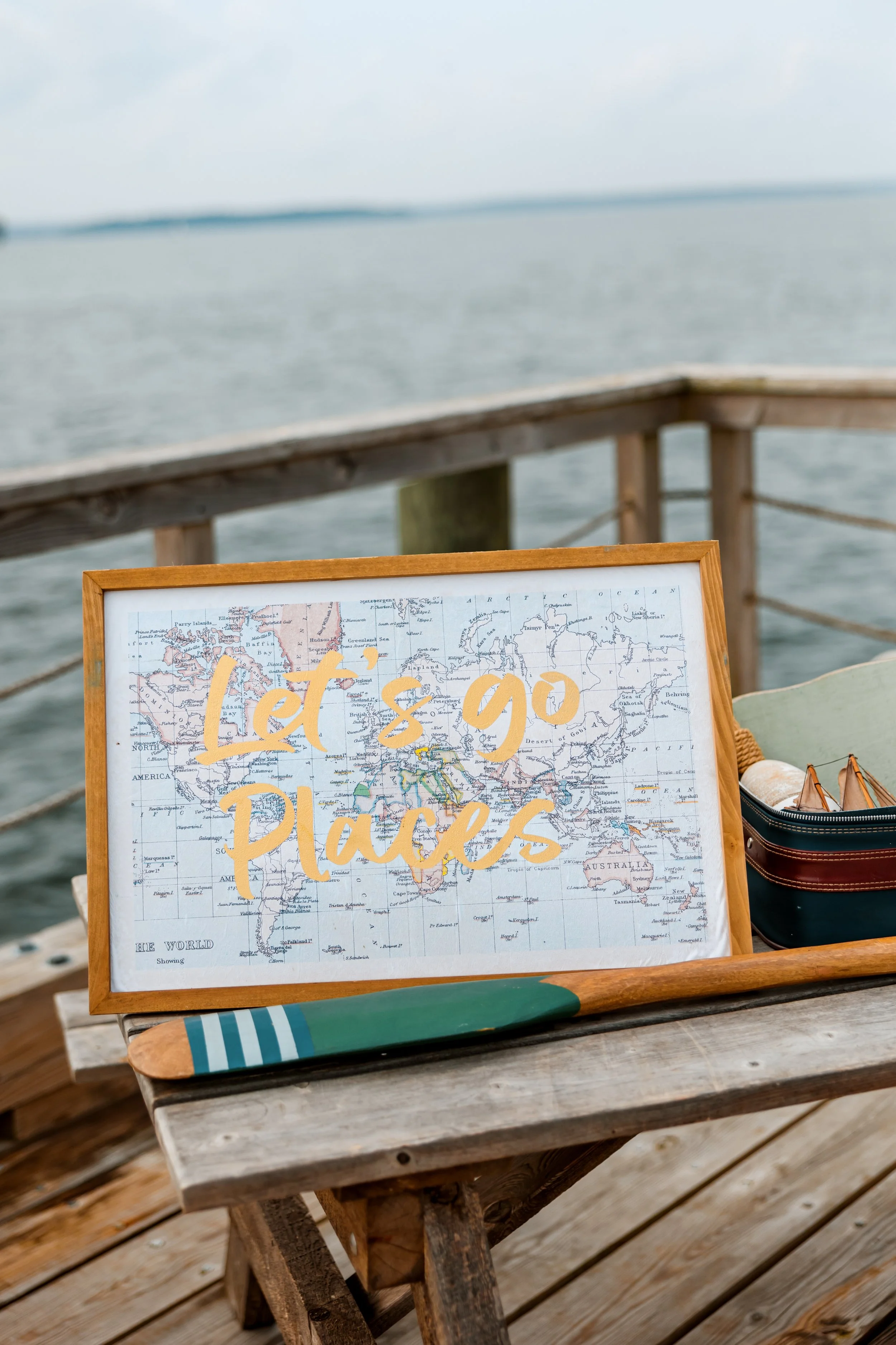 A framed map with the phrase "Let's go places" written on it in orange, placed on a wooden table near water, with a paddle and small life vests nearby.