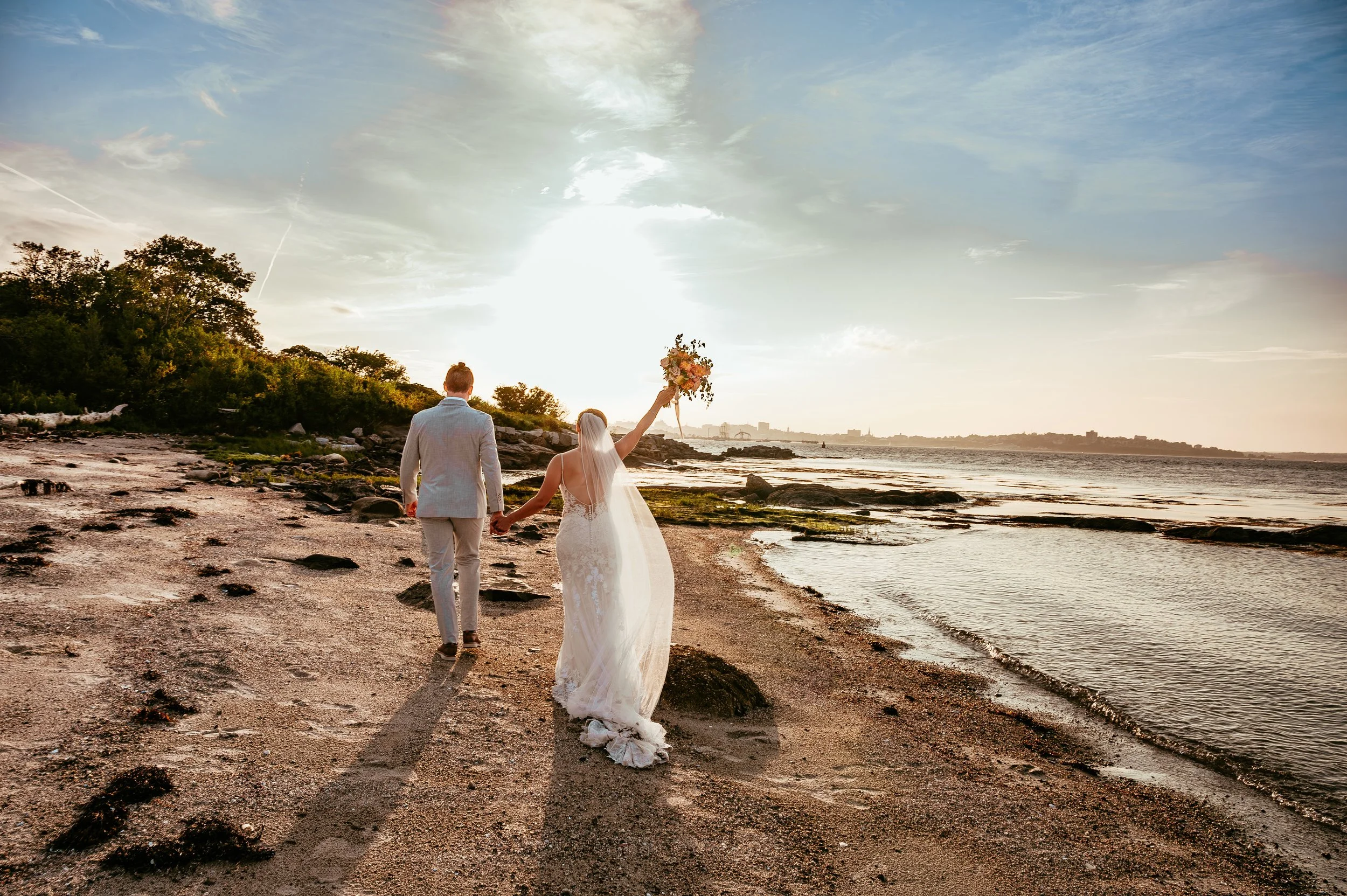 A bride and groom walking hand in hand on a sandy beach during sunset, with the bride holding a bouquet and the groom in a light gray suit.