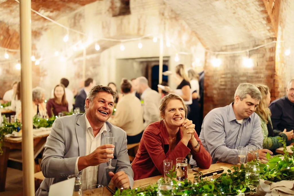 People enjoying a celebration in a rustic, brick-walled venue decorated with string lights and greenery.