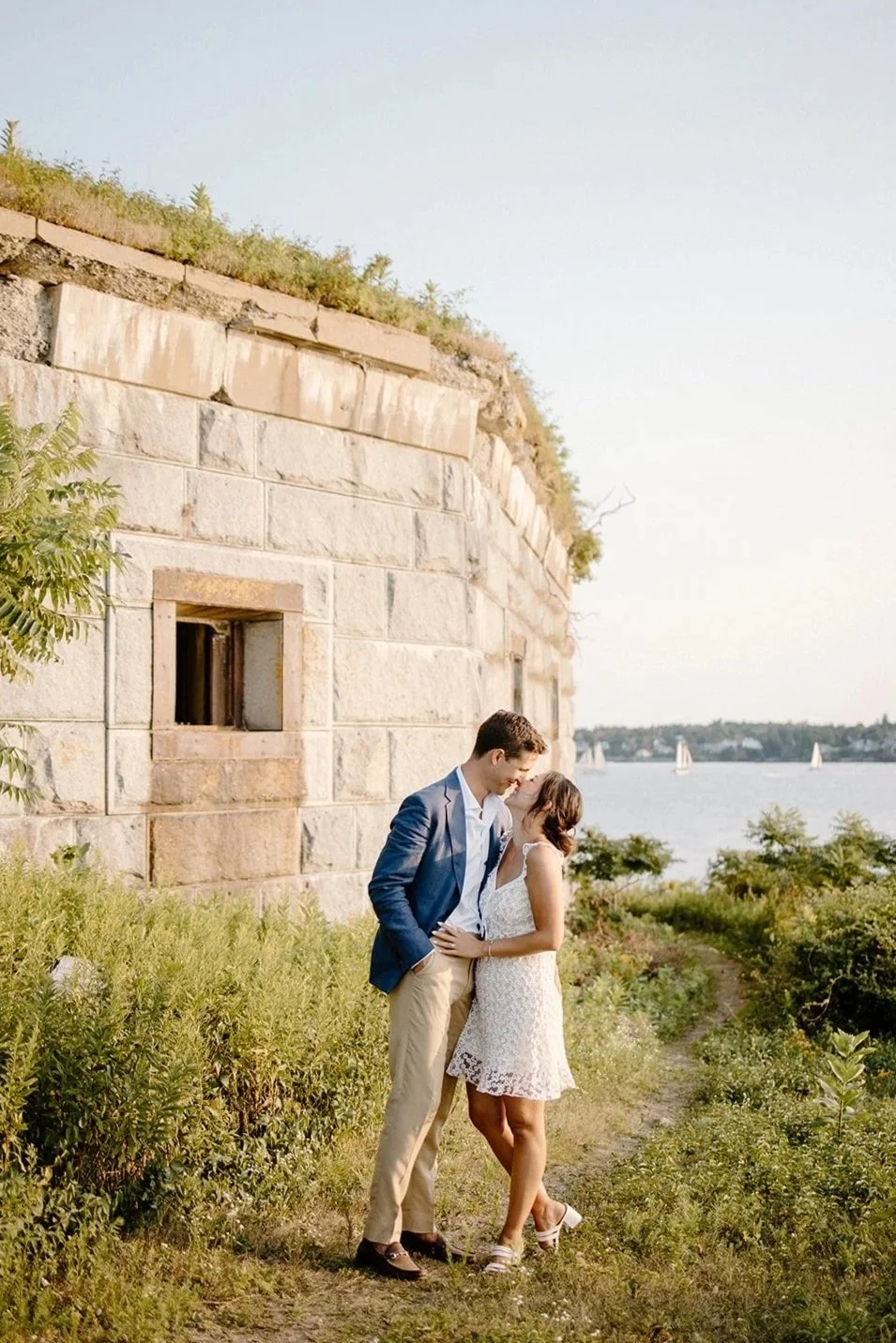 A young couple sharing a kiss outdoors near a stone wall by a body of water with sailboats in the background, during what appears to be late afternoon or early evening.