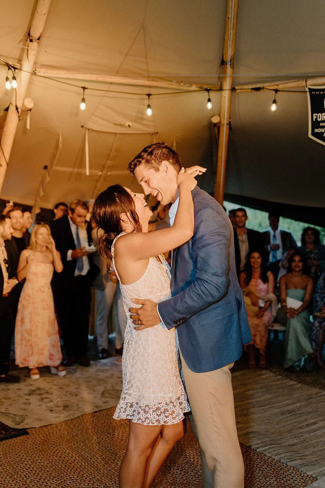 A couple shares a dance at a wedding reception under a tent, surrounded by guests watching and celebrating.