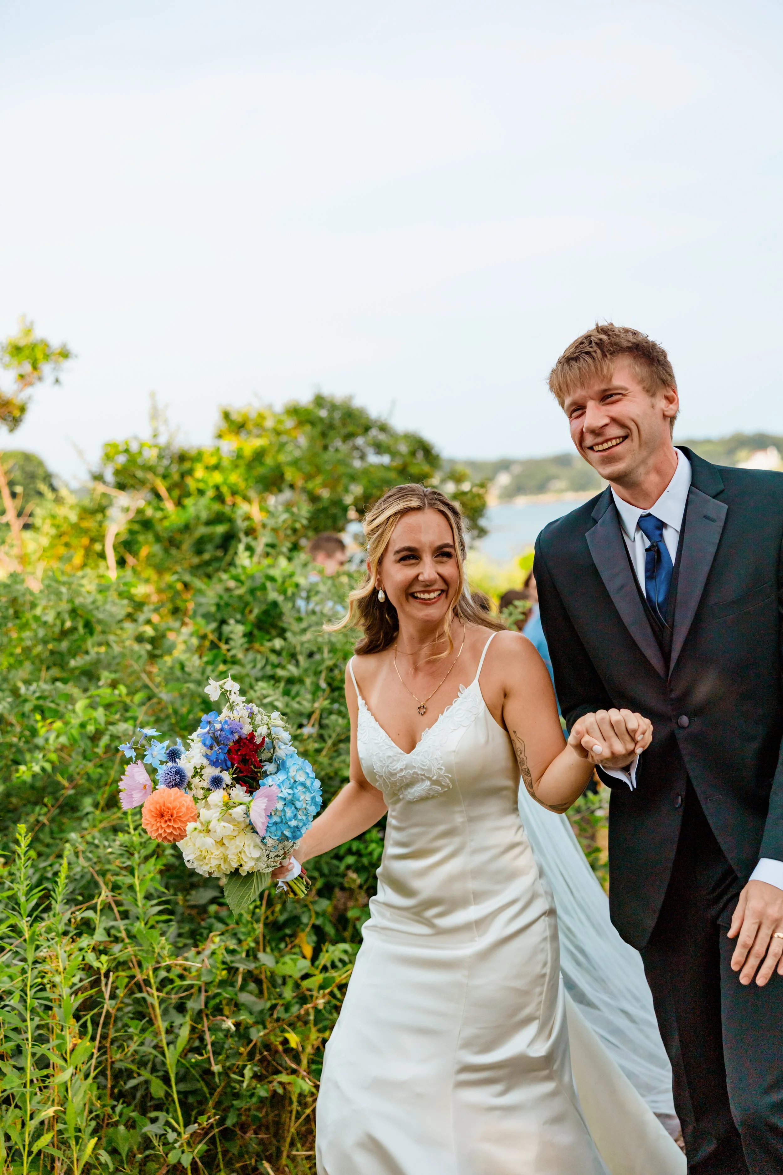 A newlywed couple is enjoying their wedding day outdoors amidst greenery; the bride is holding a colorful bouquet and smiling, while the groom is dressed in a suit, also smiling.