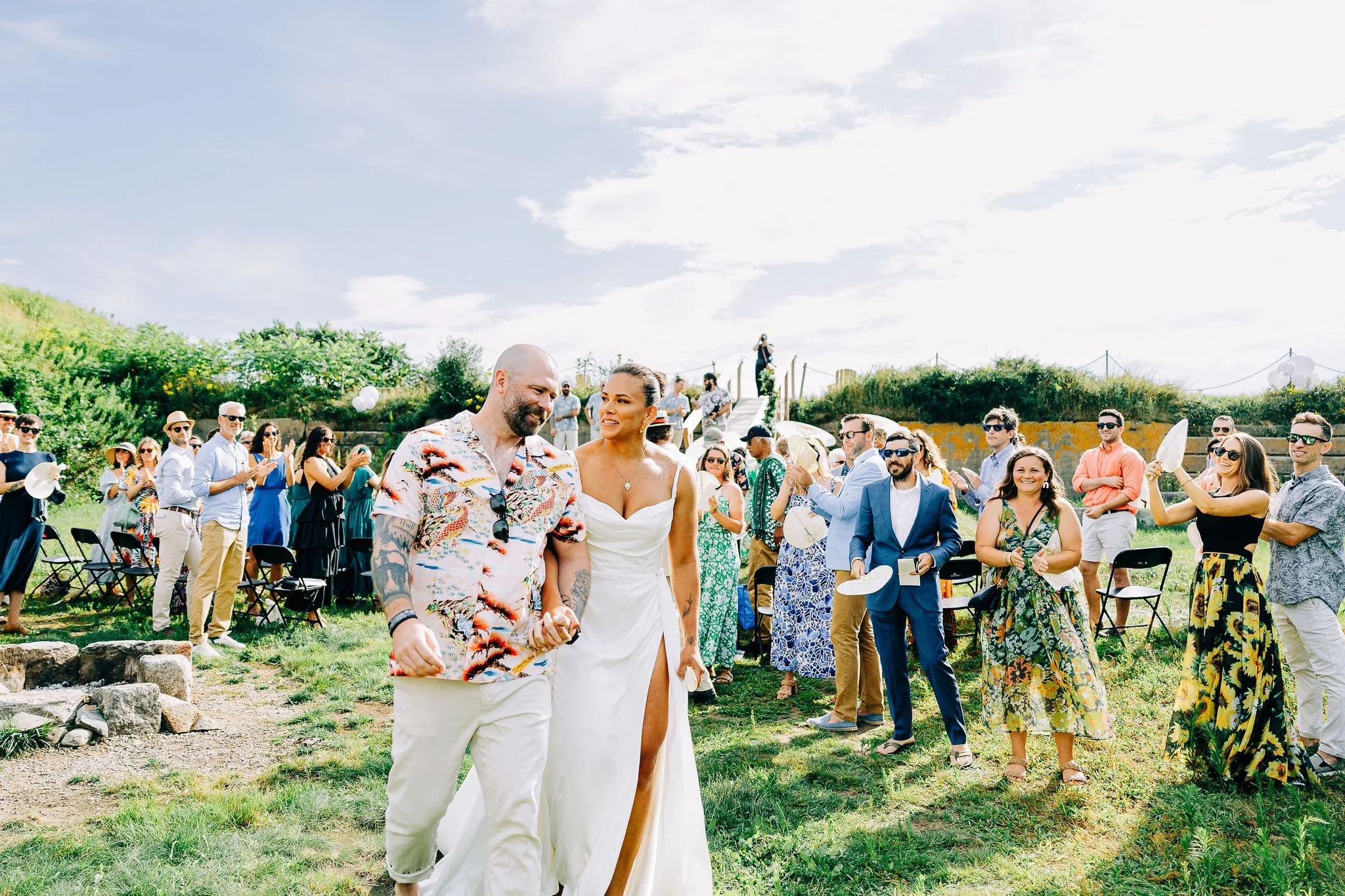 A couple is walking hand in hand at their outdoor wedding, surrounded by guests clapping and celebrating on a sunny day.