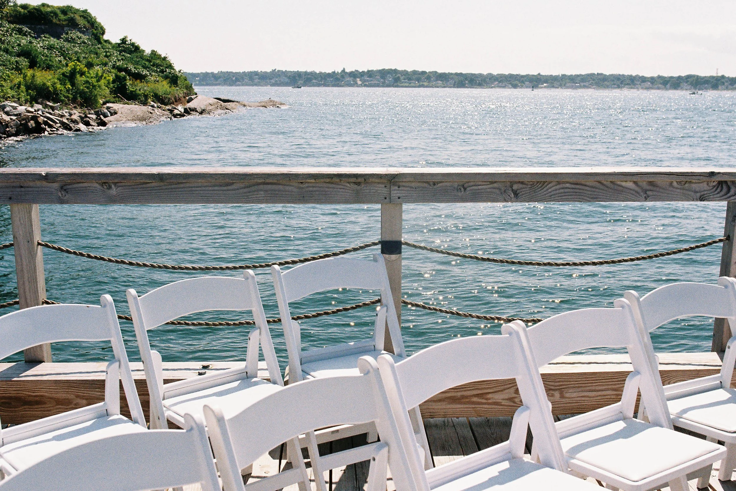 Empty white folding chairs on a wooden dock overlooking a body of water with a rocky shoreline on the left and a distant landmass under a partly cloudy sky.