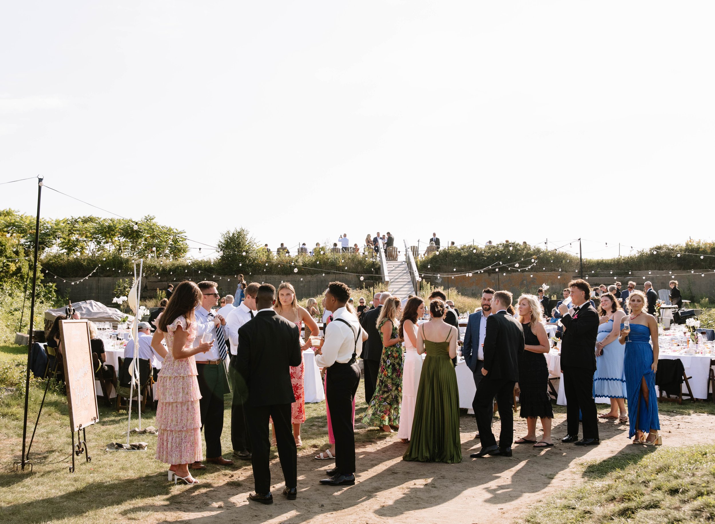 Group of people at an outdoor wedding reception, standing and talking on a grassy area with tables and string lights, under a bright sky.
