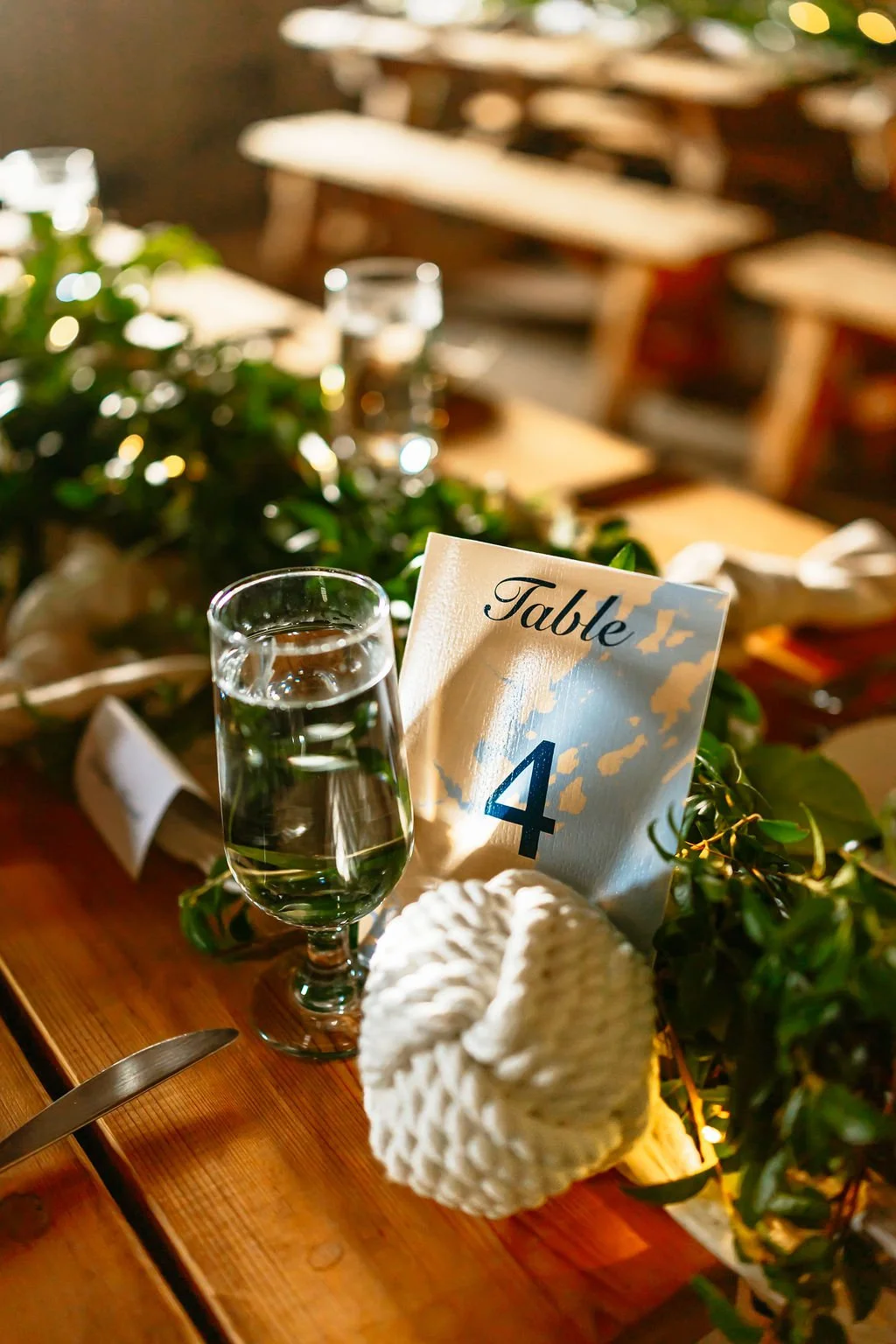 A table setting with a glass of water, a knitted white heart-shaped ornament, and a table number card labeled 'Table 4', decorated with greenery, at a warmly lit event or restaurant.