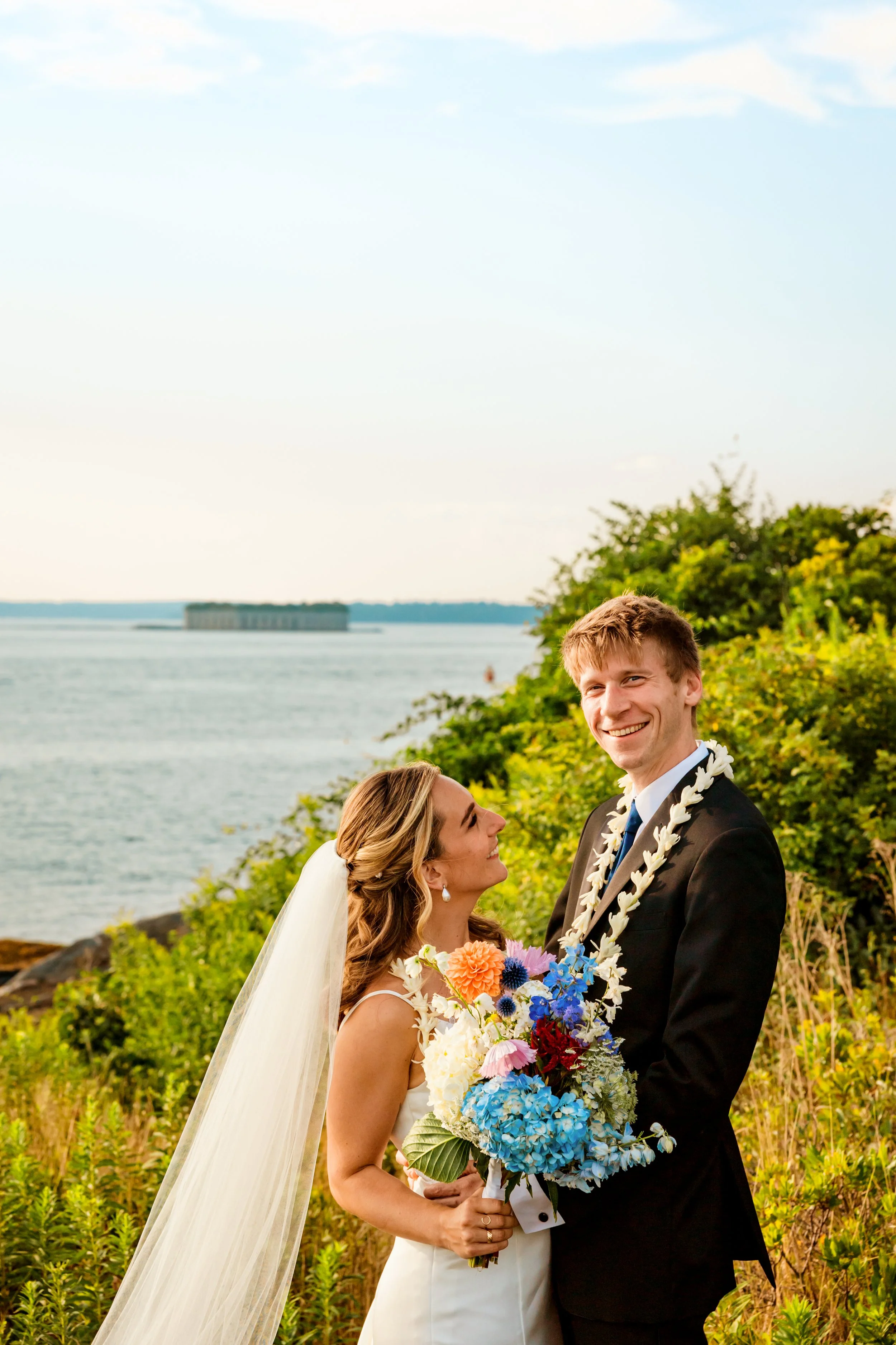 A bride and groom outdoors near water, smiling at each other, with the bride holding a large colorful bouquet, wearing a white dress and veil, and the groom in a black suit with a lei around his neck.
