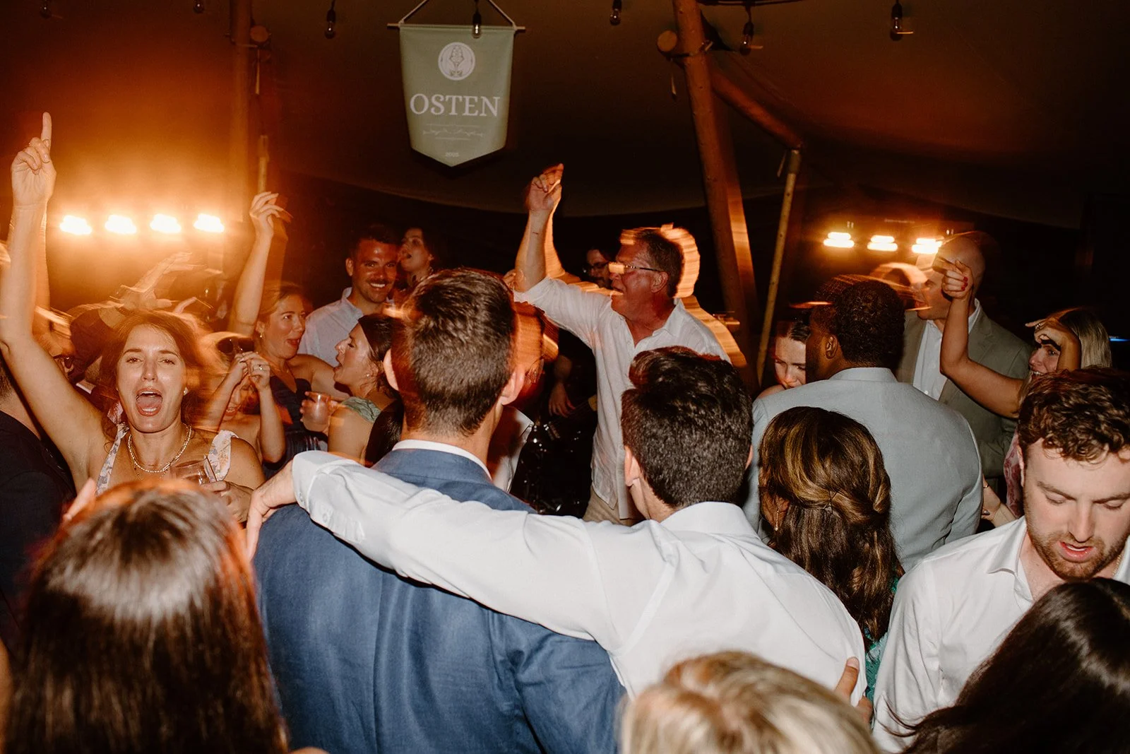 People dancing and celebrating at a party or wedding reception, with some raising their hands and smiling.