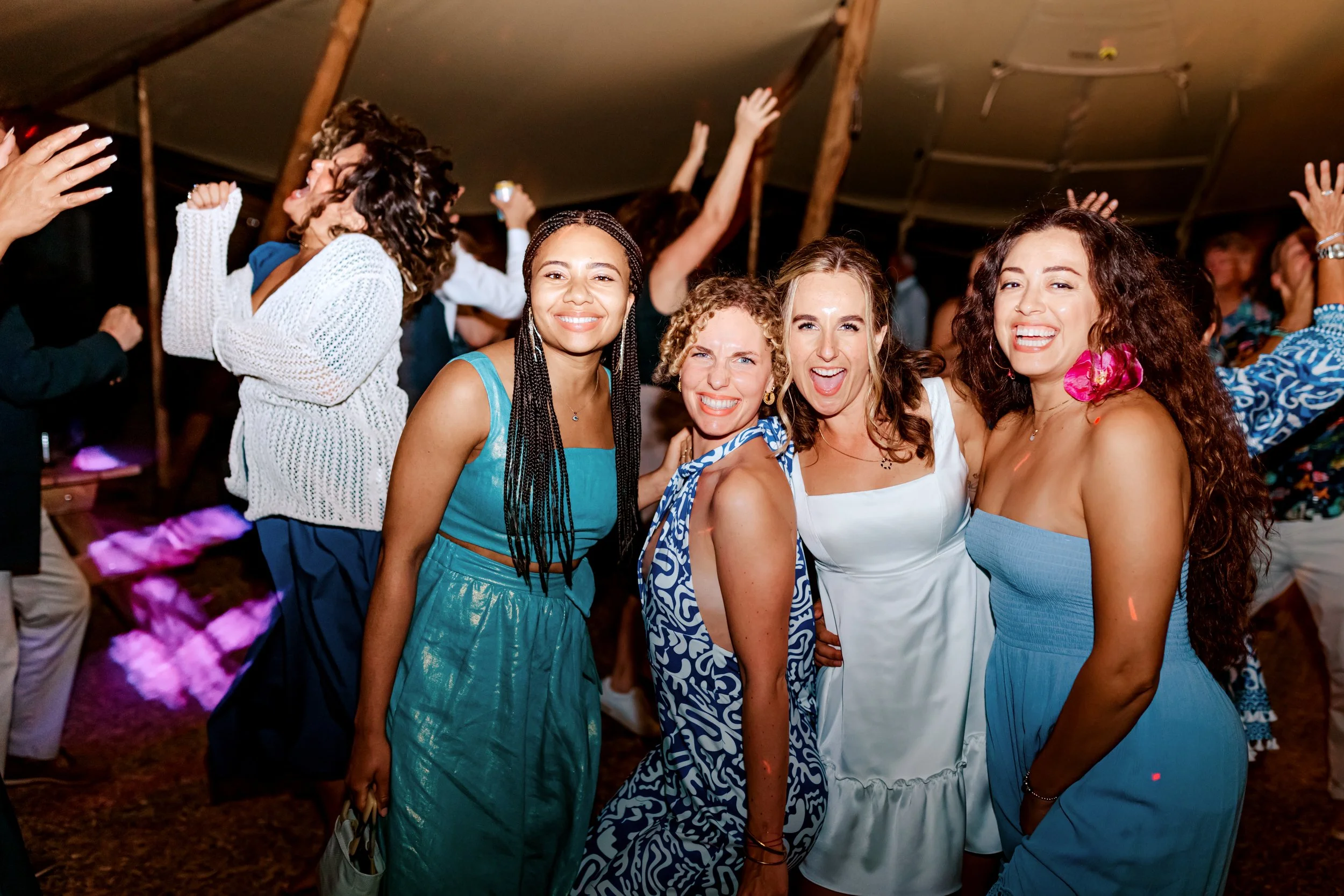 Four women in colorful dresses smiling and dancing at a lively party or celebration inside a tent, with other guests in the background.