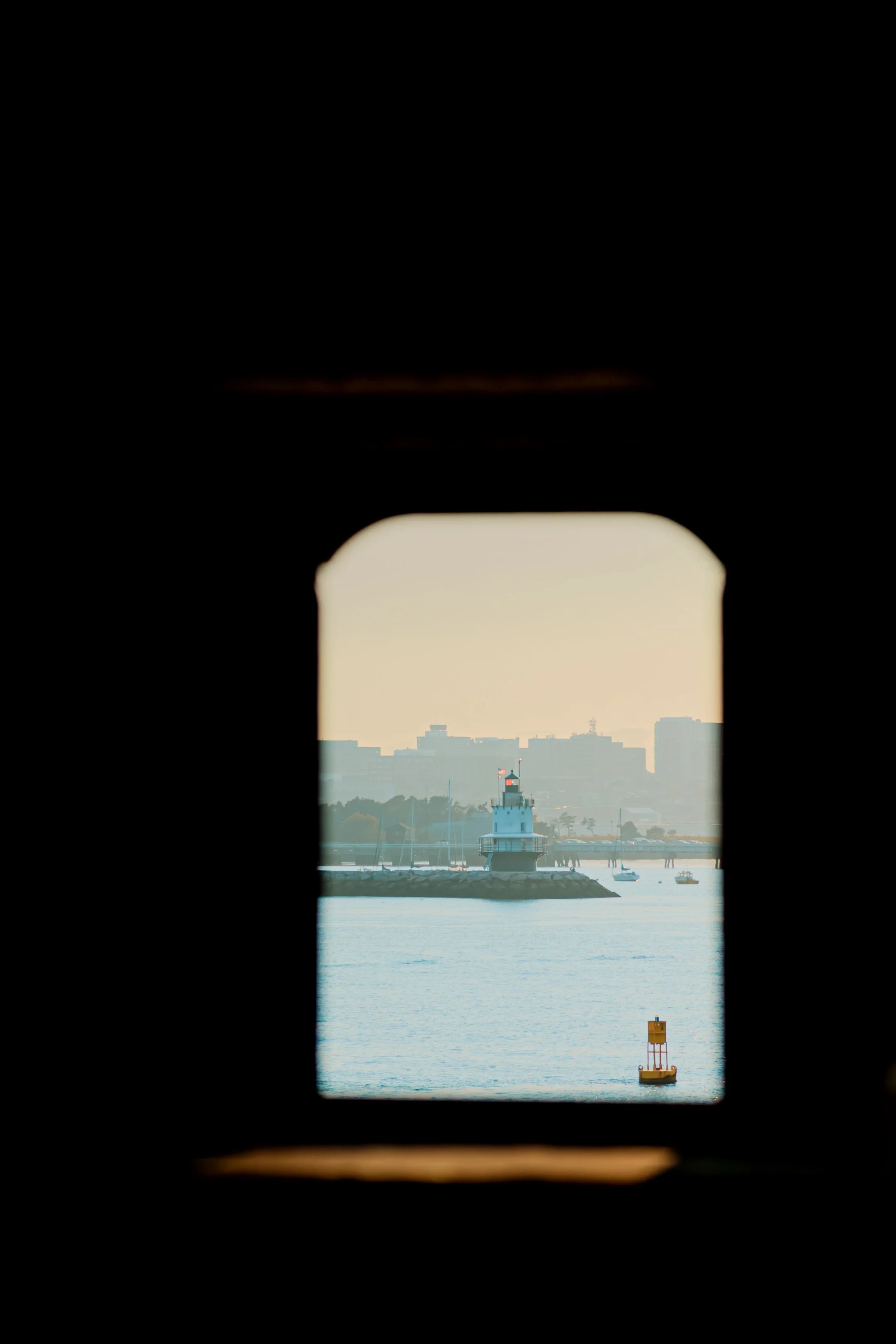 View of a harbor through a small window, featuring a lighthouse, sailboats, and a city skyline in the distance during sunset.