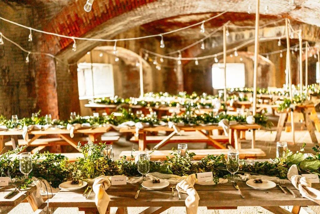 Decorated rustic banquet hall with long wooden tables, green foliage, string lights, and beige napkins set for a celebration